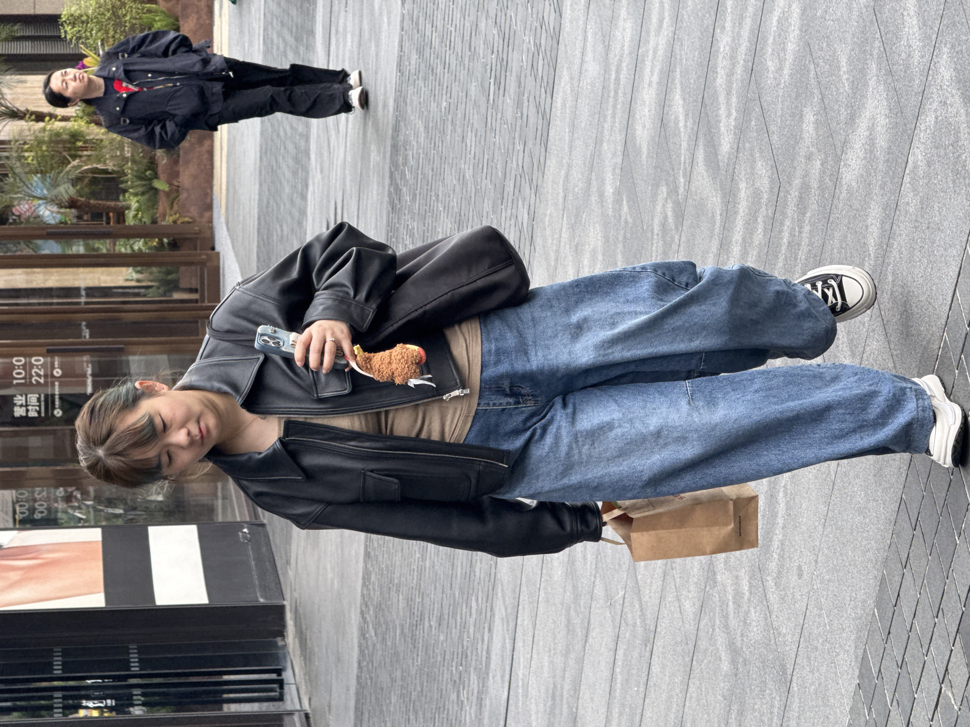 A full-length shot shows a young woman on a pedestrian walkway and another person in the background in Yuzhong District, China. The walkway appears to be made of gray brick. The woman in the foreground is facing left and is looking down and smiling. She has a leather jacket, a purse slung over her shoulder, jeans, and white sneakers. She is holding a phone and a small stuffed animal. She is also holding a brown paper shopping bag. The person in the background is wearing a dark jacket and dark pants and is walking away from the camera. They are standing on a similar walkway. Behind the woman in the foreground is a building, possibly a store, with signage in Chinese and English. Behind the person in the background are bushes.