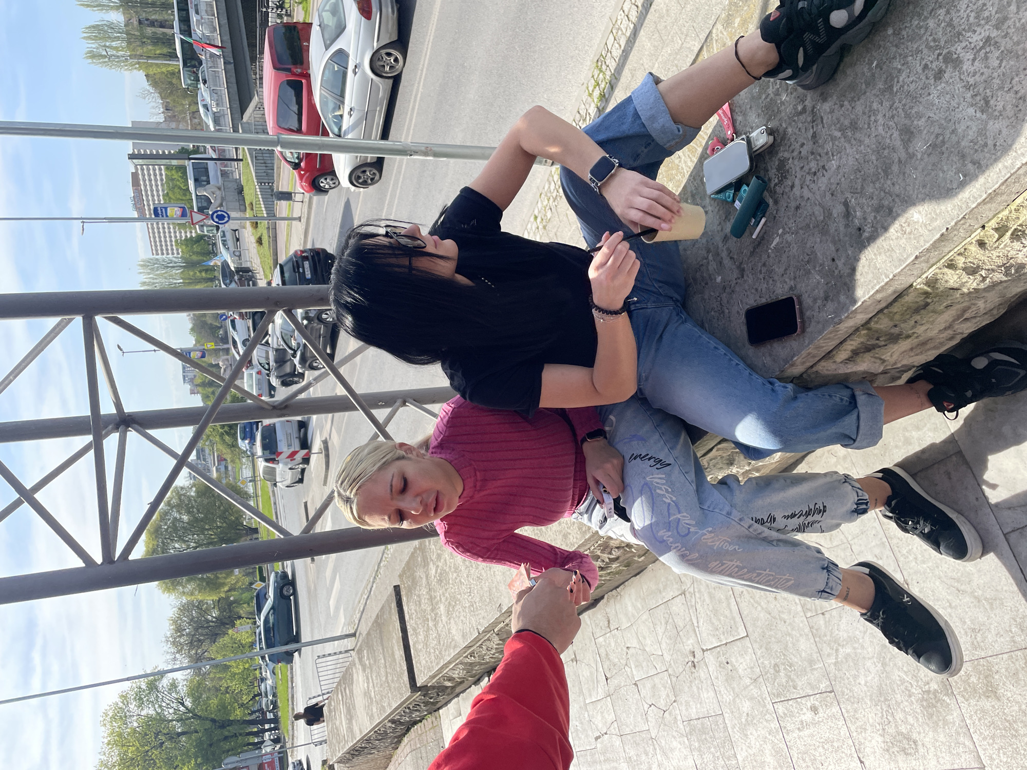 This media file captures an outdoor urban scene in Plovdiv, Bulgaria, on a sunny day. Two young women are seated on a concrete ledge, resting. In the foreground, a person's hand, wearing a red long-sleeved top, extends a banknote towards one of the women.
The woman on the left has long dark hair, wearing a black t-shirt, cuffed blue jeans, and black sneakers with white soles. She wears an Apple Watch on her left wrist and several black beaded bracelets on her right wrist, one with a small silver charm. Sunglasses are resting on her head, and she is holding a light brown cup with a straw or stirrer. Beside her on the concrete ledge are various personal items, including a black smartphone, a green and white electronic cigarette or vape device, and a silver flip-style device with a pink keychain.
The woman on the right has blonde hair, wearing a ribbed pink long-sleeved top, light-wash blue jeans, and black sneakers. Her jeans feature white graphic text, including "energy," "less stress," "perfection," and "authenticity," along with other patterns. She is looking down towards the money being offered.
The setting includes a street with parked cars in the background, a large building, and trees with green foliage under a clear blue sky. Metal railings are visible in the immediate foreground, suggesting an elevated viewpoint for the shot. A white bus with what appears to be a Bulgarian flag is visible further down the street. Road signs, including a blue circular sign with a white arrow, are also present in the distance. The activity suggests a casual social interaction, possibly an exchange of money for a purchase or a personal transaction.