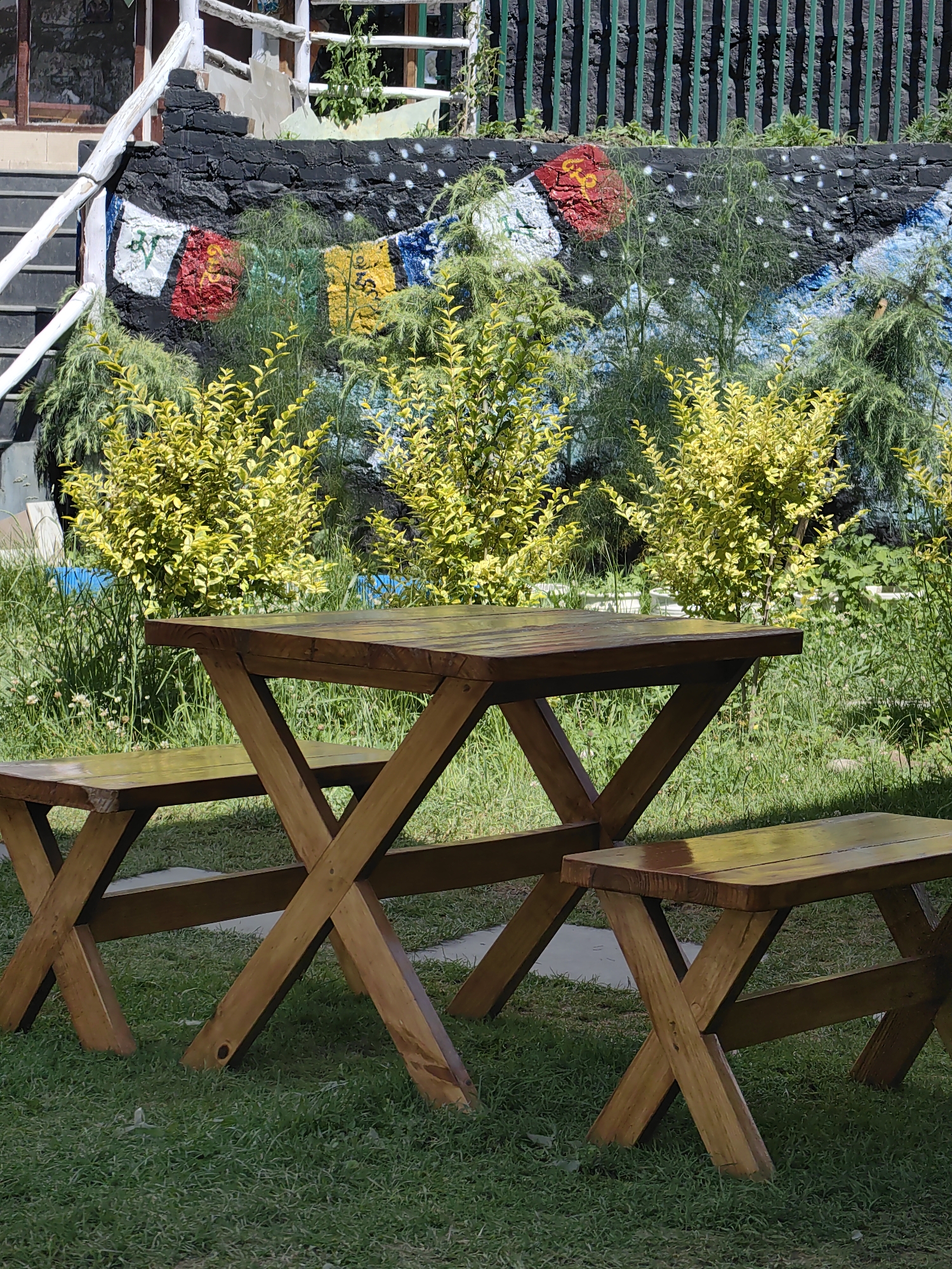 This image depicts a wooden picnic table and two benches situated on a grassy area in Vashisht, India. Behind the picnic set, there is a black stone wall adorned with colorful graffiti, featuring abstract shapes and what appear to be prayer flags. Lush green and yellow foliage grows in front of the wall, partially obscuring it. The background includes a glimpse of a building with stairs and a white railing, as well as a green metal fence. The overall setting suggests an outdoor recreational space.