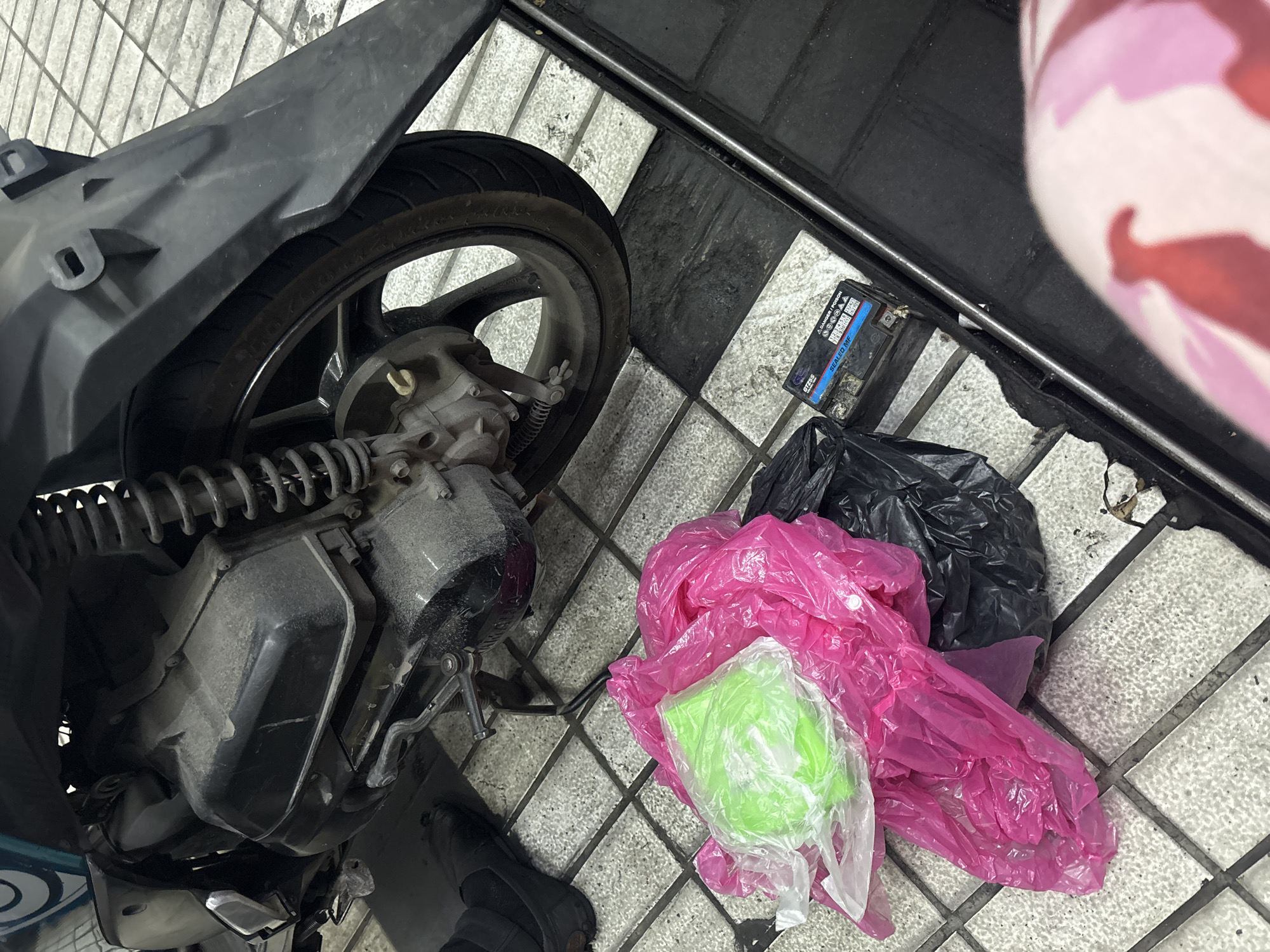 In North Jakarta, Indonesia, a partially visible motorcycle with a dusty engine and rear wheel is parked on a tiled floor. In front of the motorcycle, a pile of plastic bags, one bright green and others pink and black, are present on the ground. A black battery, possibly for the motorcycle, sits next to the bags. A person's lower body, clad in pink and white camouflage-patterned leggings, is visible in the bottom right corner of the image. The scene appears to be outdoors or in a well-lit, open area with the tiled floor extending throughout.