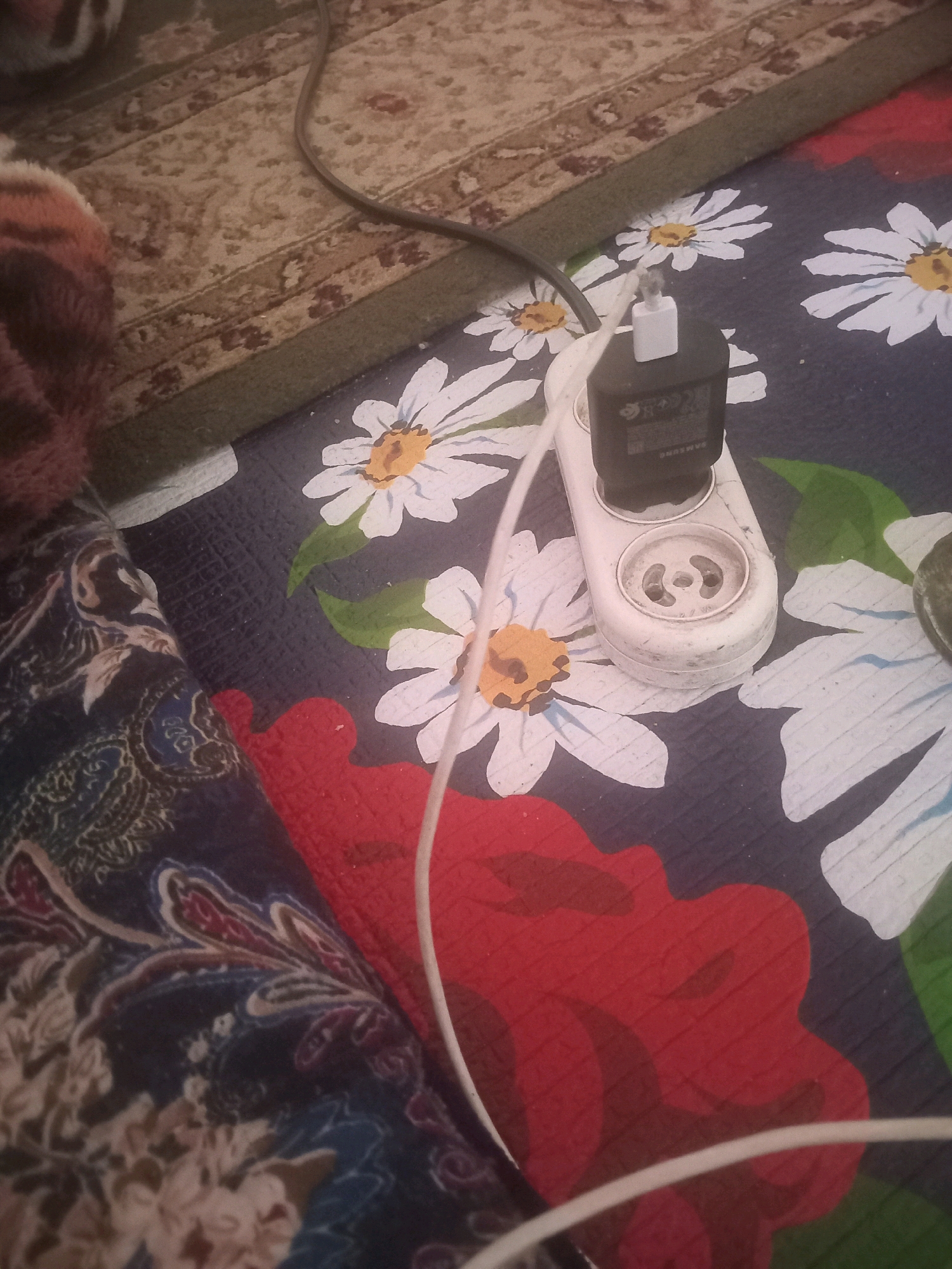 The image shows a white electrical outlet strip with a black Samsung phone charger plugged into it. The outlet strip is on a dark blue surface with a floral pattern of white daisies and red flowers. A white cable is plugged into the outlet strip and extends out of the frame. The scene appears to be indoors, likely a room with carpeting and textiles. There are no people visible in the image. The location is not specified in the image.