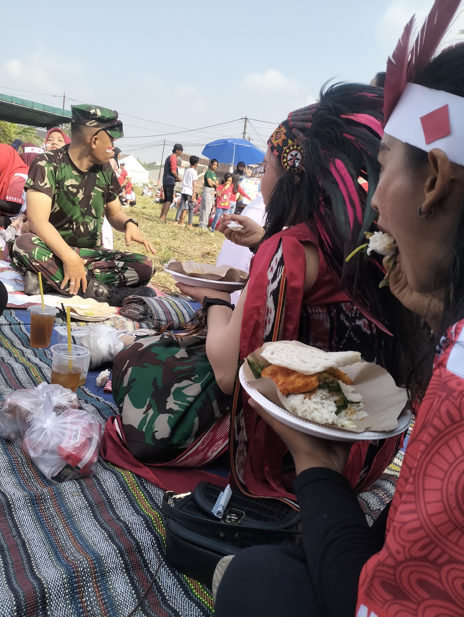 In Jetis, Indonesia, on a grassy field under a clear sky, a group of people are gathered for an outdoor event. In the foreground, a man in camouflage military uniform sits on a colorful striped mat, looking to his left. To his right, a woman wearing an elaborate headdress with colorful feathers and beads, and a red and black traditional garment, holds a plate of food. Another woman, with a similar headdress and a red garment with intricate patterns, is in the process of eating from a plate of rice, vegetables, and fried food. Several plastic cups with drinks, plastic bags containing food items, and a black purse are scattered on the mat. In the background, more people are visible, some seated and others standing, with a blue umbrella providing shade in the distance. The scene appears to be a festive gathering or celebration.