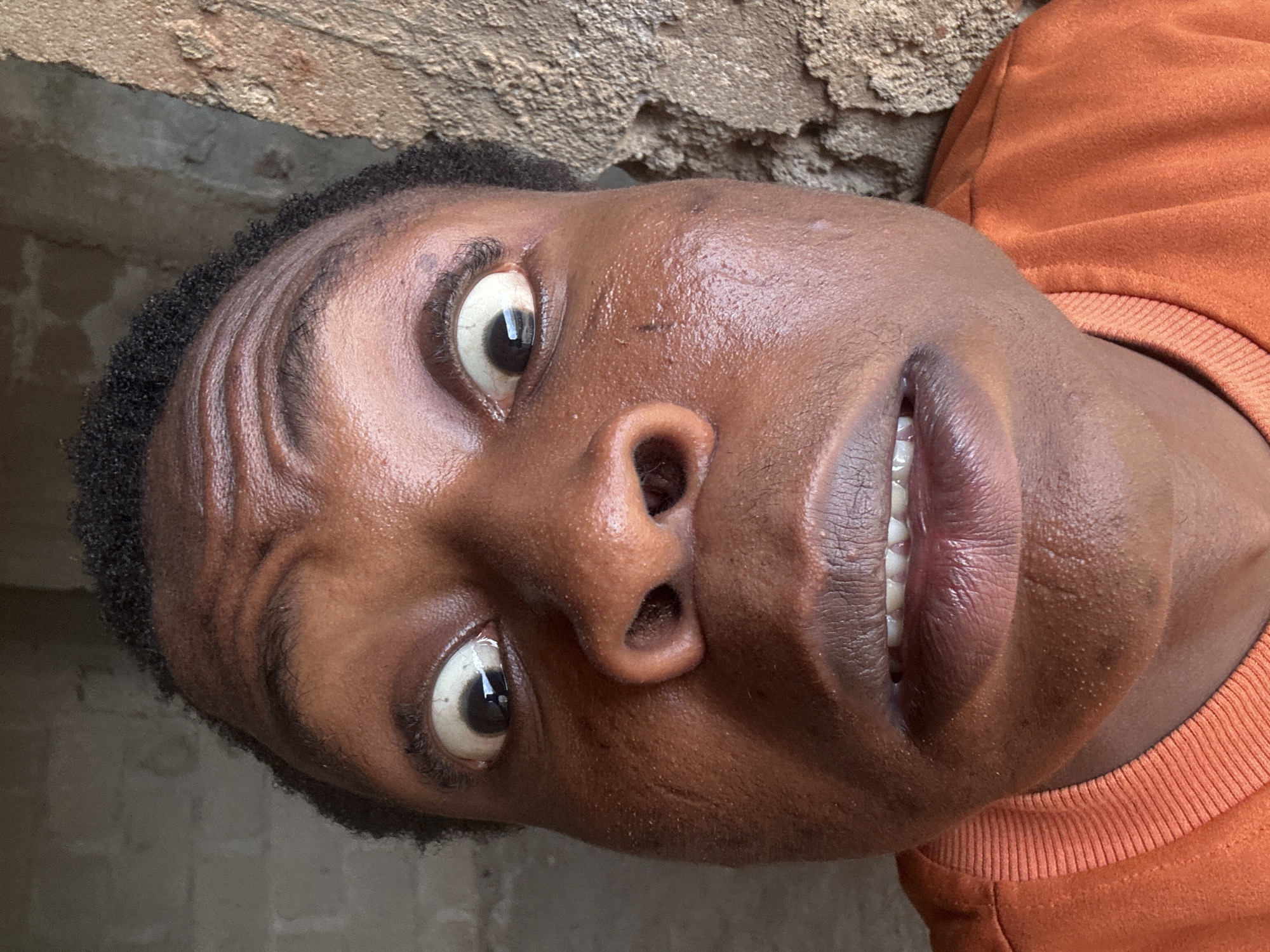 The image shows a close-up of a person's face, tilted at an angle. The person has dark skin, dark hair, and white teeth visible through slightly parted lips. The person's eyes are wide and looking upwards. They are wearing an orange sweatshirt. The background consists of a rough, textured wall, possibly made of concrete. The location is Unknown, Unknown.
