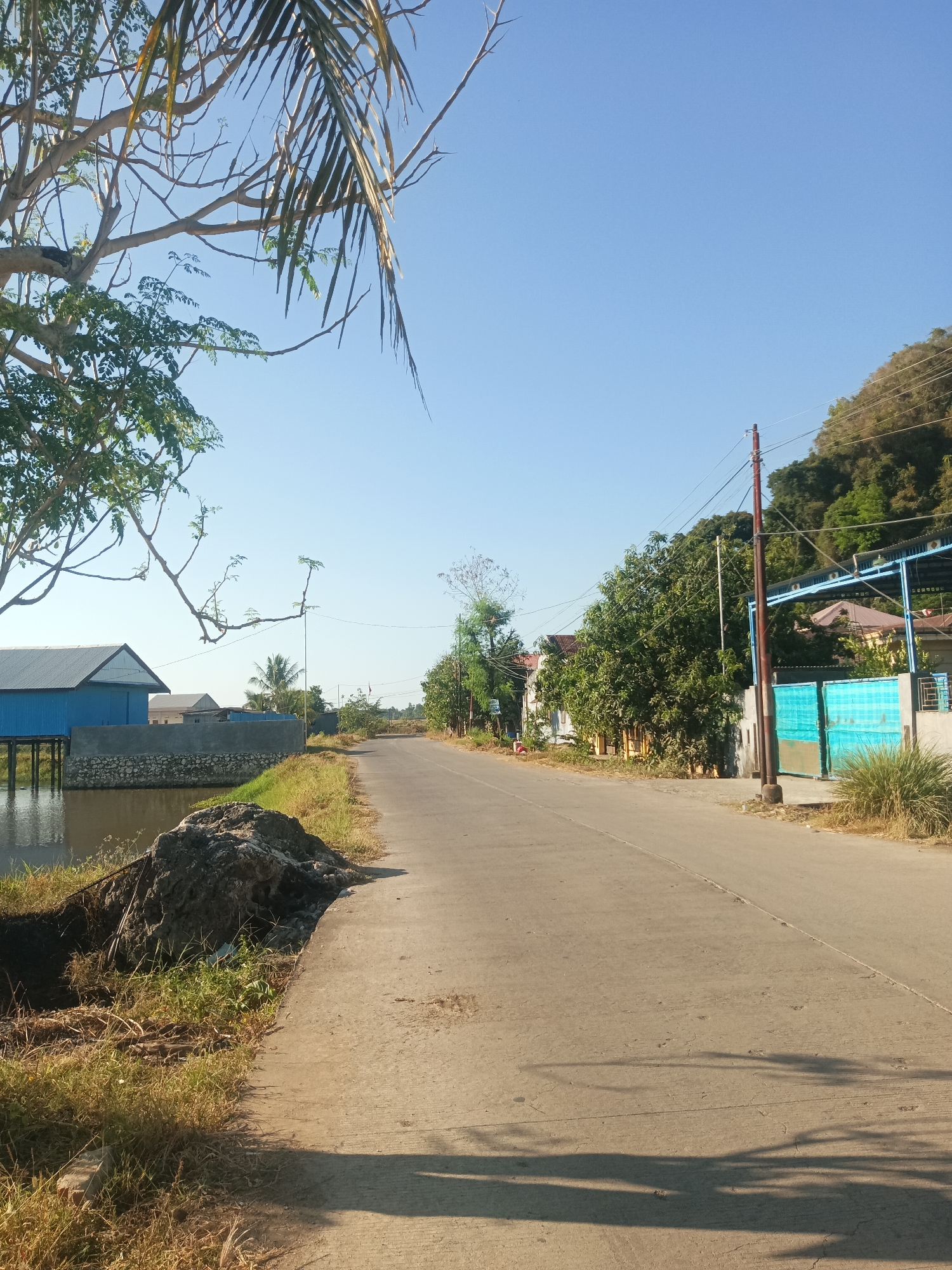 The image depicts a rural road in Bowong Cindea, Indonesia, on a clear, sunny day. A paved road runs through the center of the frame, curving gently towards the horizon. To the left of the road, a body of water is visible, with a blue building on stilts situated beside it. The building has a gray stone foundation and a corrugated metal roof. Adjacent to the water and the building is a pile of dirt and debris. Further down the road, on the right side, are several houses and trees, with a steep, wooded hill in the background. A utility pole with overhead wires stands near the right side of the road. The sky is a clear, vibrant blue, and shadows from trees and branches are cast across the road, indicating the time of day is likely mid-morning or afternoon. The overall scene conveys a sense of peaceful, rural life.