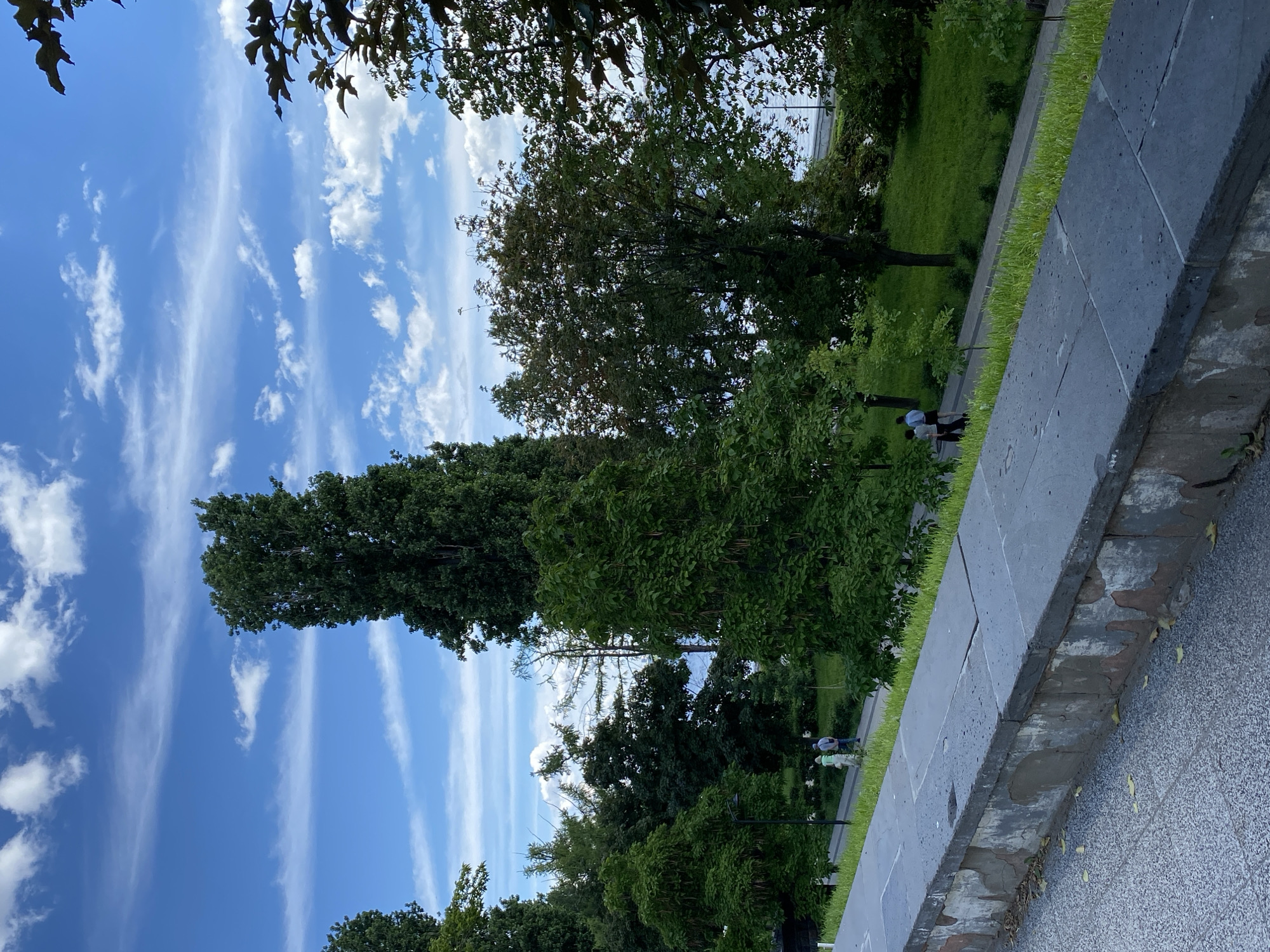 The image shows a park scene with a blue sky filled with wispy white clouds. Tall, lush green trees are prominent throughout the frame, with some foliage in the foreground suggesting dappled sunlight. In the middle ground, a paved pathway runs alongside a grassy area. A few people are visible walking along the pathway, appearing to enjoy a leisurely stroll. A stone wall or elevated border is in the foreground, constructed from large, rectangular gray stones. The overall impression is a peaceful and pleasant outdoor setting, likely during the daytime given the bright sky. The orientation of the image is portrait, with the sky at the top and the ground at the bottom.
