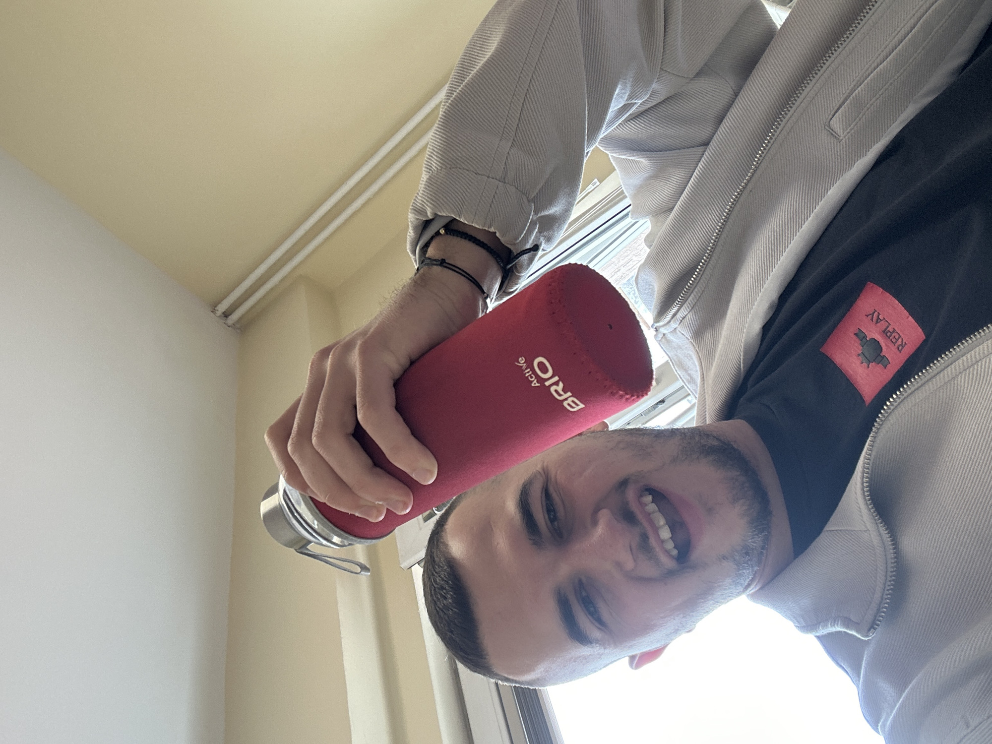 A male individual is captured in a close-up, upward-angled selfie, smiling broadly with an open mouth and visible teeth. He has short dark hair and stubble, and is wearing a light grey, possibly corduroy, zip-up jacket over a dark t-shirt. A red rectangular patch with "REPLAY" in white text and a logo is visible on the lower right of his t-shirt. Several thin black bracelets adorn his right wrist. He holds a red bottle with a silver metallic top and a loop in his raised right hand; white text "Activ BRIO" is present on the bottle's red sleeve. The background indicates an indoor setting in Plovdiv, Bulgaria, with light beige walls and a white ceiling. A bright window is visible on the left, and two horizontal white pipes run along the upper right wall.