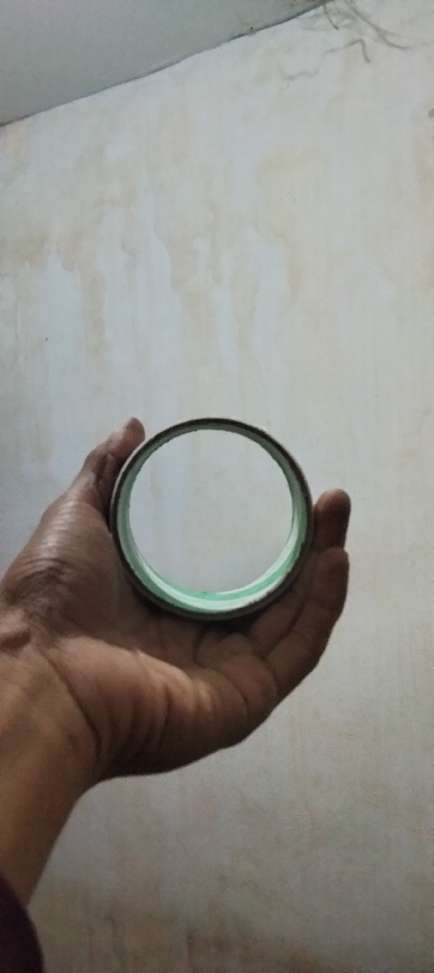 A close-up shot shows a hand holding a circular object with a clear center and a green rim against a mottled, off-white wall. The object appears to be a magnifying glass or a lens. The hand is dark-skinned with visible wrinkles. The wall has water stains and discoloration. The scene is likely indoors, given the wall texture and lighting. The image does not provide any visual information that would suggest it is located in Antirogo, Indonesia.