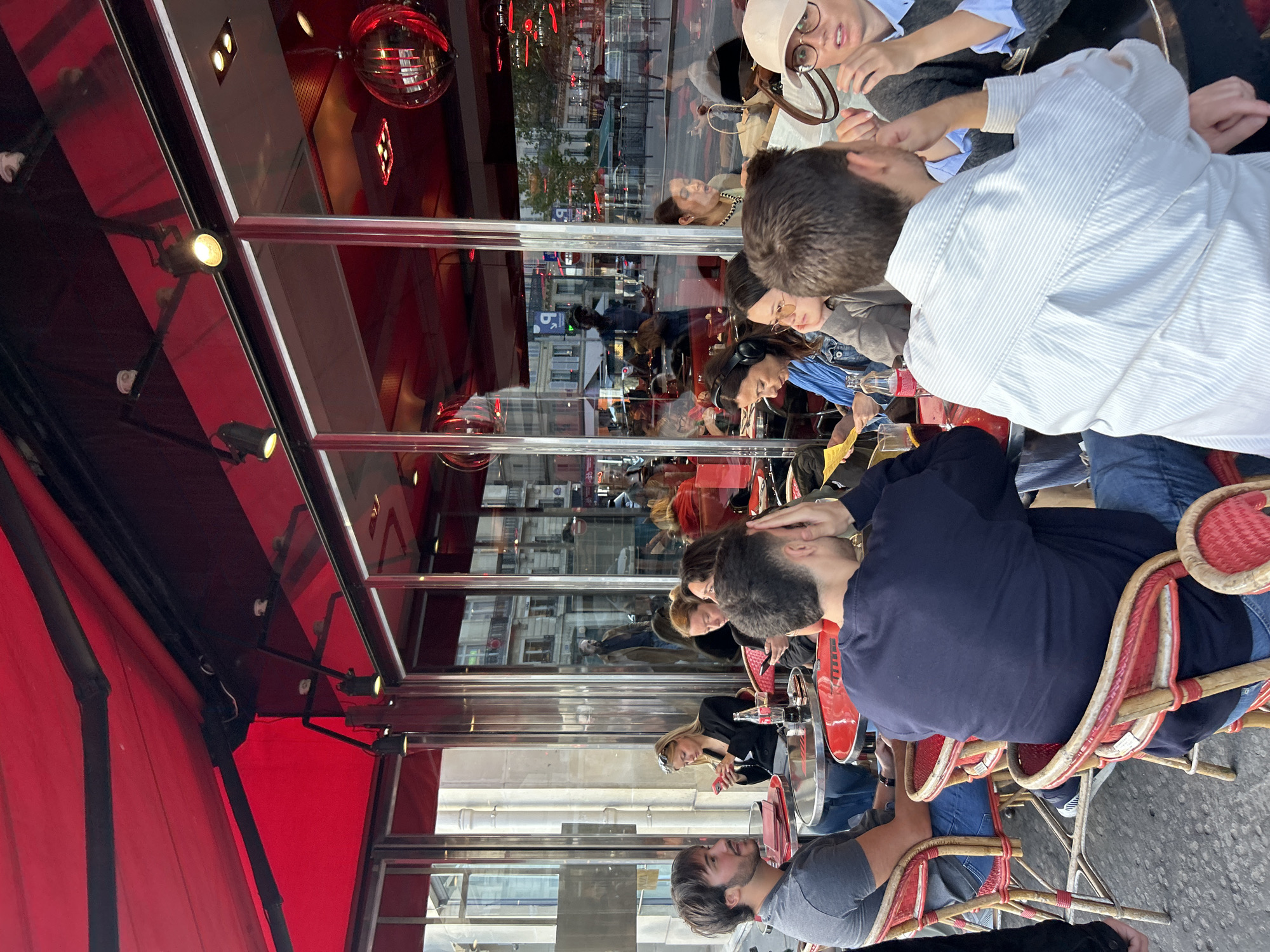 A group of people are seated at tables outside a Parisian cafe, under a red awning. The cafe's interior is visible through large glass windows, with reflections of street scenes and buildings. The people are casually dressed and appear to be engaged in conversation or using their phones. Some tables have drinks and menus on them. The atmosphere suggests a typical day at an outdoor cafe in Paris.