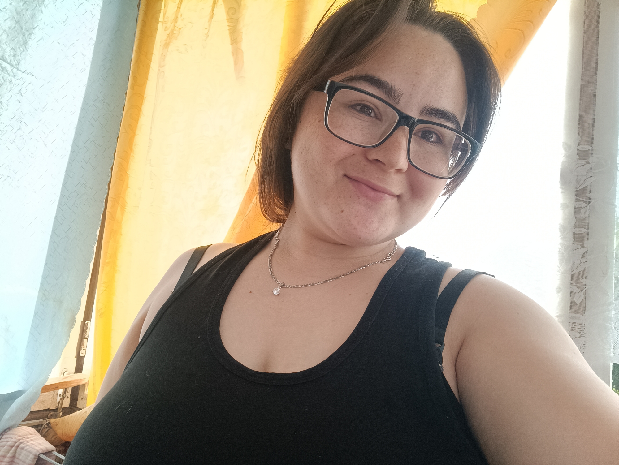 A woman is taking a selfie indoors, with curtains in the background. She is wearing glasses and a black tank top, and a necklace with a diamond pendant. The location is not specified in the image.