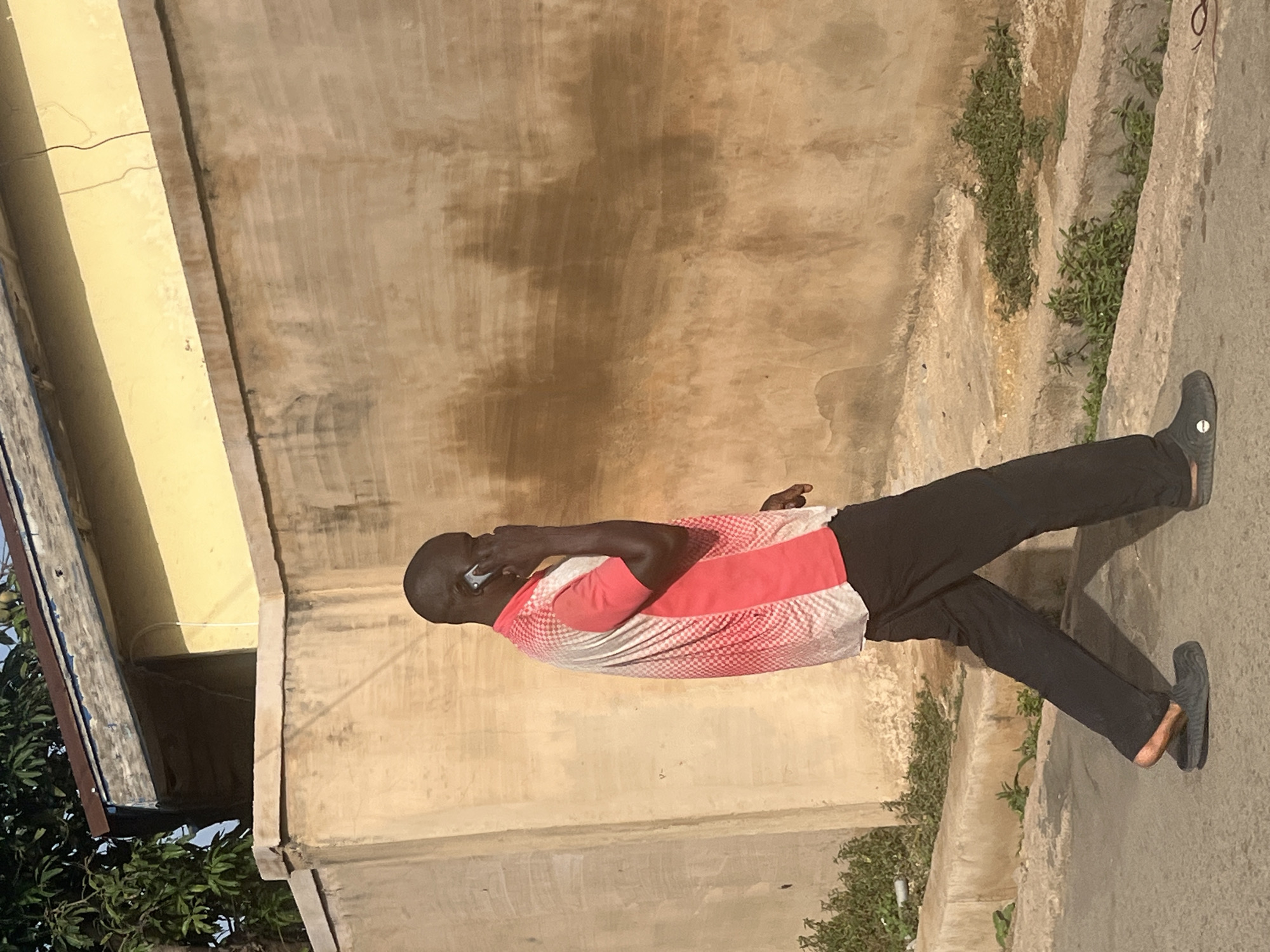 A man is walking on a paved surface next to a concrete wall while talking on a mobile phone. The man is wearing black trousers, black slip-on shoes, and a white and red shirt. The wall appears aged with a textured surface. Green vegetation is growing near the edge of the pavement. The presence of foliage suggests an outdoor location. It is not possible to determine the location in the city of Jos, Nigeria, with only the image.