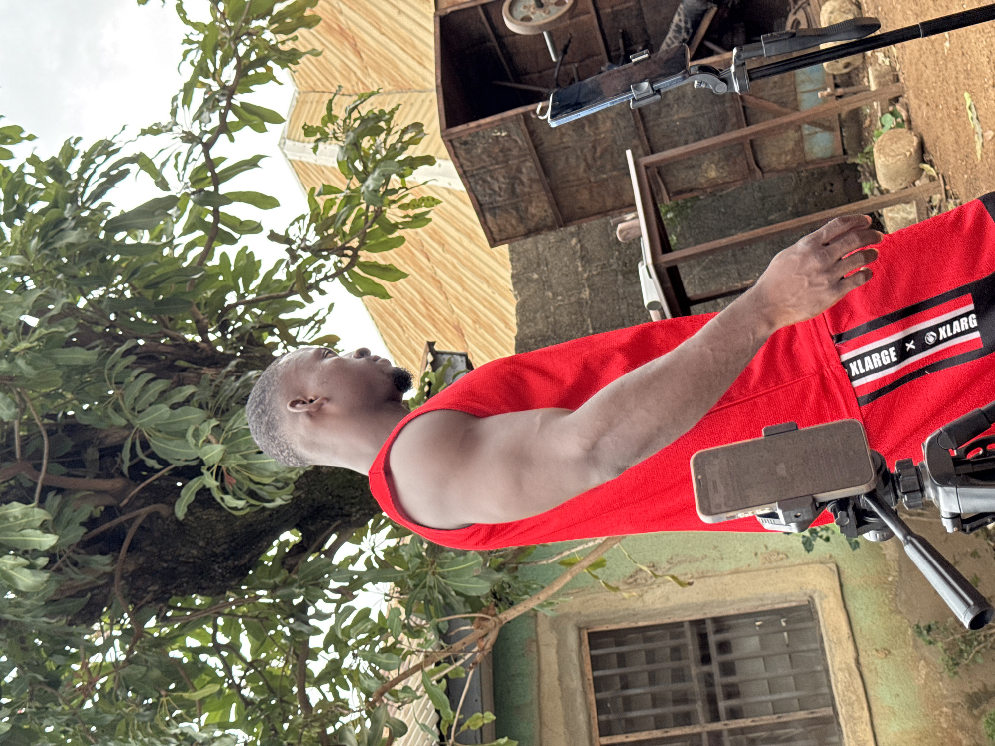 A young Black man with short hair and a goatee is standing outdoors, facing right and looking down. He is wearing a red sleeveless shirt and red shorts with white and black stripes and logos on the sides. His right arm is bent at the elbow, and his hand is resting on his hip. A cell phone mounted on a tripod is positioned in front of him, to his left.
Behind the man, a large tree with green leaves dominates the left side of the frame. To the right of the tree, a building with a corrugated metal roof is visible. In the foreground, the ground is covered with dirt and small stones. Several large, rough stones are scattered around. Behind the tripod, a rusty metal structure with a weight on a bar is partially visible. A wooden table with items on it is also in the background. The overall scene appears to be in a rural or semi-urban setting, possibly in Akwanga, Nigeria.