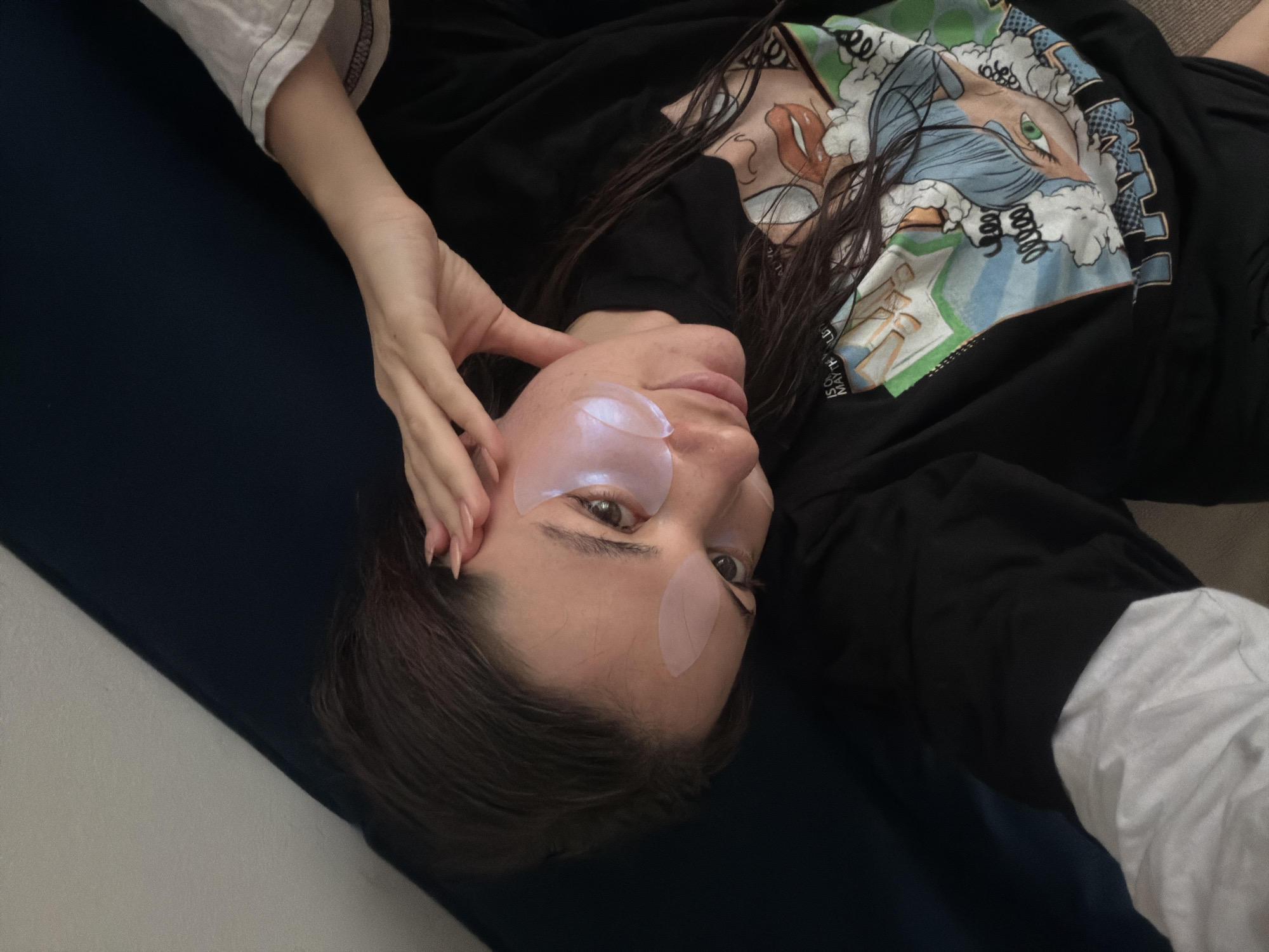 A woman lies on a dark blue fabric surface, possibly a couch or bed, wearing a black, graphic-print sweatshirt. She has fair skin, dark hair, and is wearing under-eye patches. Her right hand is near her face, with long, manicured fingernails. A white sleeve is visible near her left arm. The background includes a plain, white surface. The location is assumed to be indoors, potentially a home or apartment. The location of Belgrade, Serbia, cannot be verified.