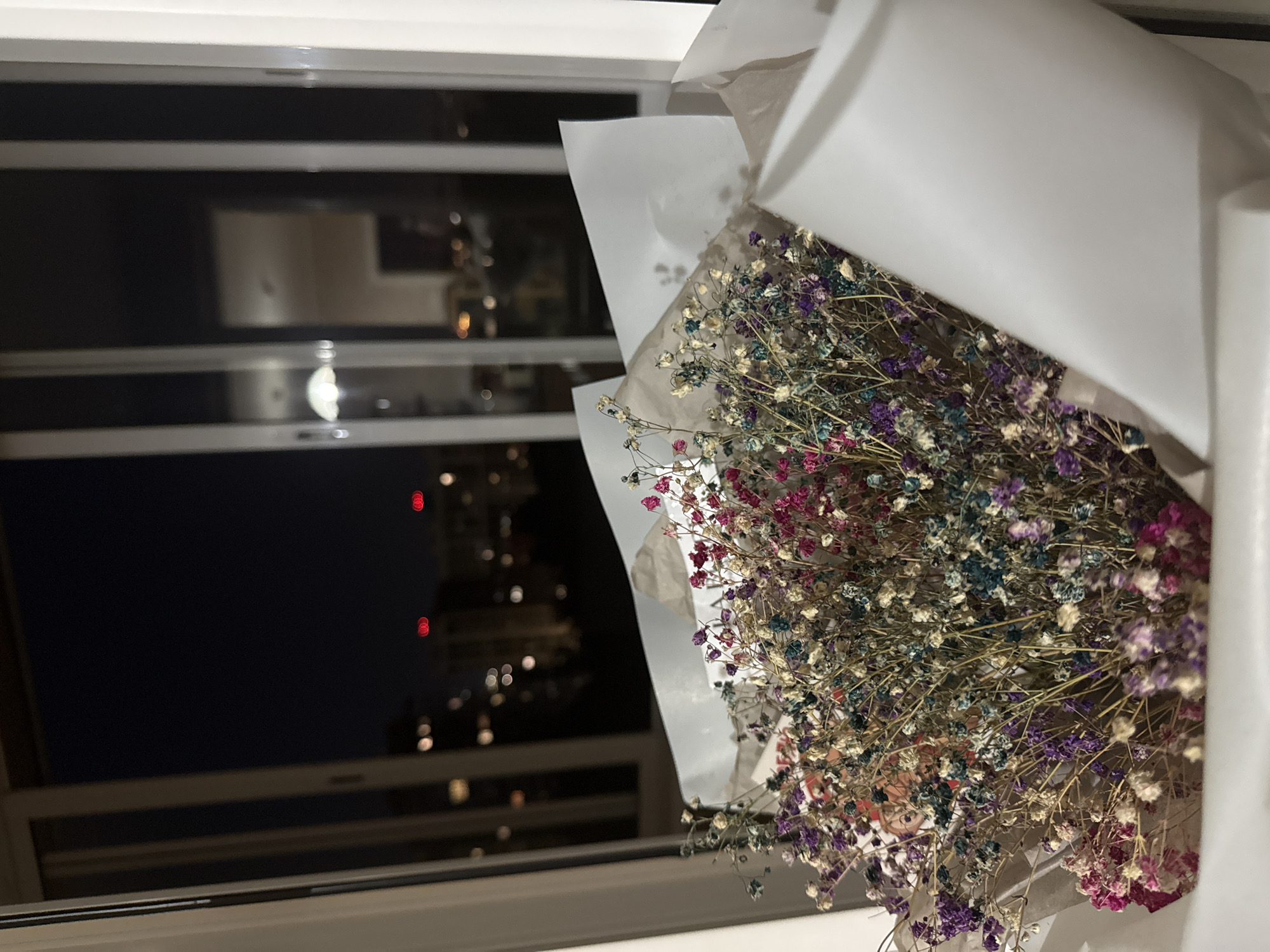 A close-up, low-angle shot shows a bouquet of dried baby's breath flowers with a cityscape visible through a window at night. The flowers are a mix of white, pink, purple, and blue. They are wrapped in white and brown paper. The background features a dark night sky with blurred city lights and buildings. The reflection in the window suggests an indoor setting.
