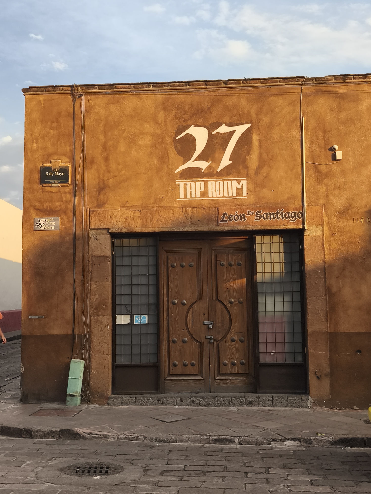 The image captures the facade of a building in Querétaro, Mexico. The structure is a warm, brown-orange stucco. Above the dark wooden double doors are the words "TAP ROOM" and the number "27" in white lettering, along with the words "León de Santiago". To the left of the entrance, a sign reads "5 de Mayo" (Fifth of May). A green electrical box is near the base of the wall. The scene is illuminated by sunlight. The building is on a street made of cobblestones.