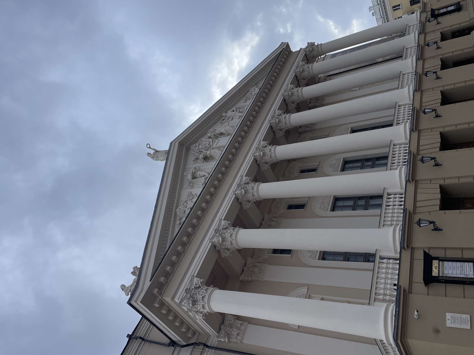 The image presents a low-angle view of a grand classical-style building under an overcast sky, with the building extending upwards from the bottom and angled slightly to the left. The prominent feature is a large, triangular pediment, extensively adorned with intricate bas-relief sculptures depicting numerous figures and narrative scenes. At the apex of the pediment, a white statue of a winged figure, possibly holding a wreath, is visible. Another decorative sculpture, resembling a gargoyle or an architectural figure, can be seen at the lower left corner of the pediment.
The building's facade is supported by a series of tall, white, fluted columns, each crowned with elaborate Corinthian capitals. These columns stand in front of recessed sections of the building, which feature multiple windows, some with decorative balustrades beneath them.
To the right of this classical structure, an adjacent building with a lighter, plainer facade is partially visible. This building has several visible windows and is adorned with multiple black, antique-style wall lamps, spaced vertically. A large, black rectangular sign with white text, resembling a directory or information board, is mounted on this wall, but the text is too blurry to be legible. Below this sign, a smaller white plaque with black text and a security camera are also affixed to the wall; the text on the plaque is likewise unreadable.
The sky is heavily clouded, featuring shades of grey and white, indicating an overcast daytime. There is no discernible activity taking place. The overall impression is one of historical grandeur and architectural sophistication, characteristic of a significant public or cultural institution.