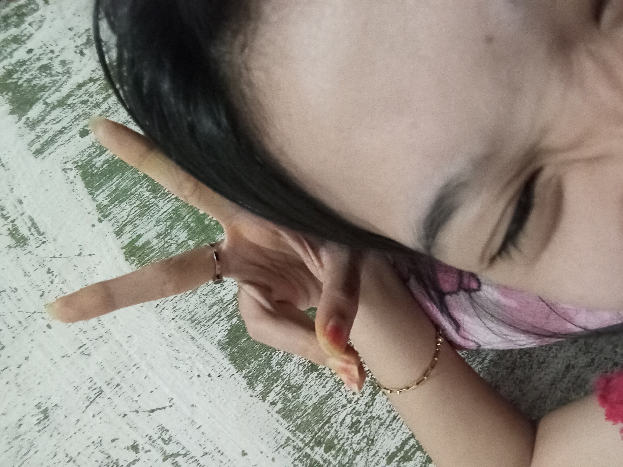 A close-up, low-angle shot shows a person's face and hand resting on a textured, light-colored surface with green painted accents. The person's left eye is partially visible, with dark eyebrows and eyelashes. Their right hand is raised, forming a peace sign with the index and middle fingers extended. A thin, gold bracelet is on their left wrist, and a small, silver ring is on the ring finger of their right hand. A hint of red fabric is visible on the upper left of the frame. The image is underexposed and has a grainy texture. The scene is set in Rejosari, Indonesia.