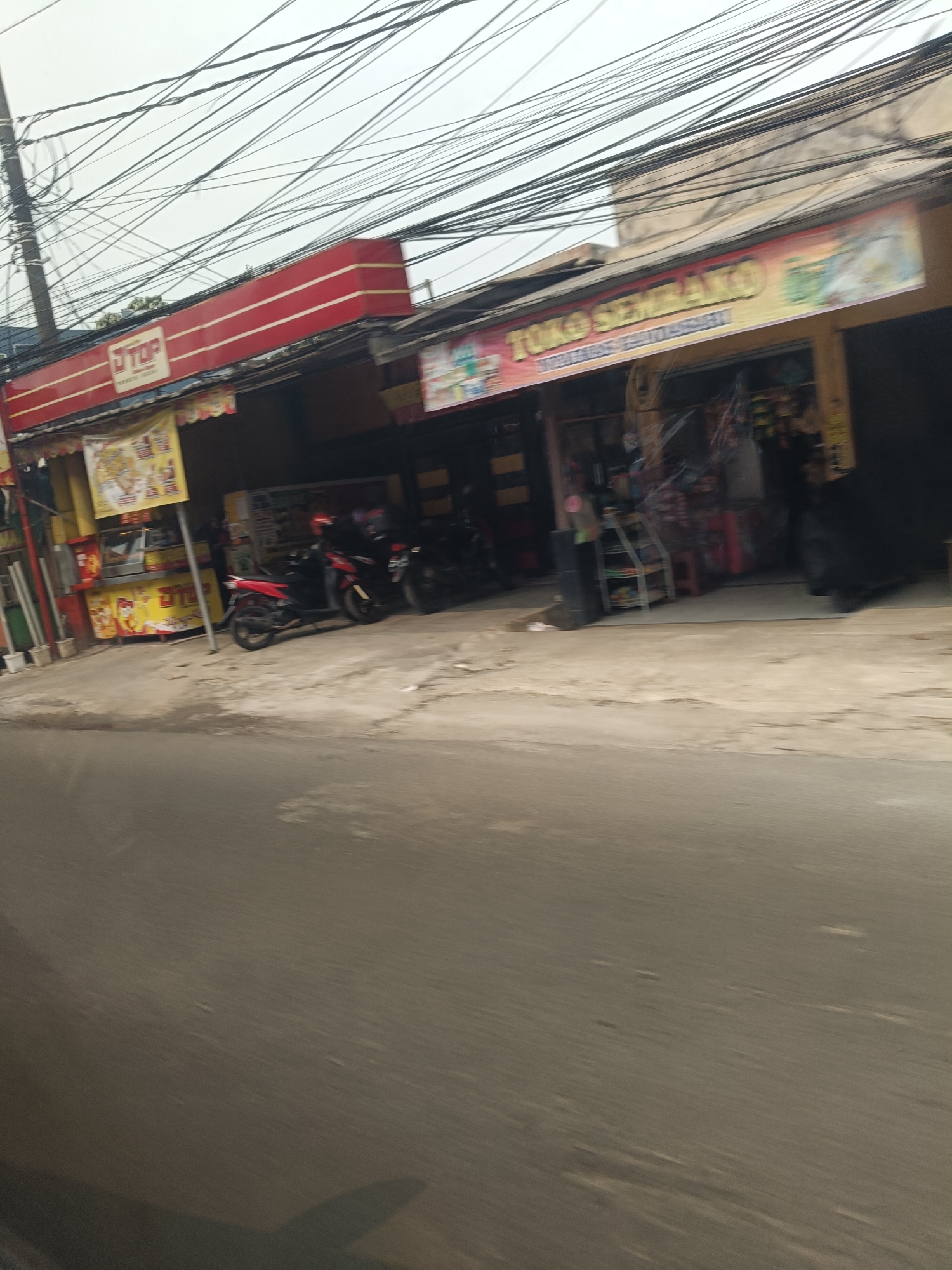 The image depicts a street scene in Saruni, Indonesia, with a clear view of two storefronts. The storefront on the left, marked with a red and white "TOP" sign, appears to be a convenience store. Next to it, a yellow food stall is visible. The storefront on the right is labeled "TOKO SEMARTO" in a banner above the entrance, suggesting it is also a shop. Several motorcycles are parked in front of the establishments, indicating typical street activity. The environment is characterized by overhead electrical wires, a paved road in the foreground, and a sidewalk or pathway in front of the shops. The overall impression is a snapshot of a busy commercial area in an Indonesian city.
