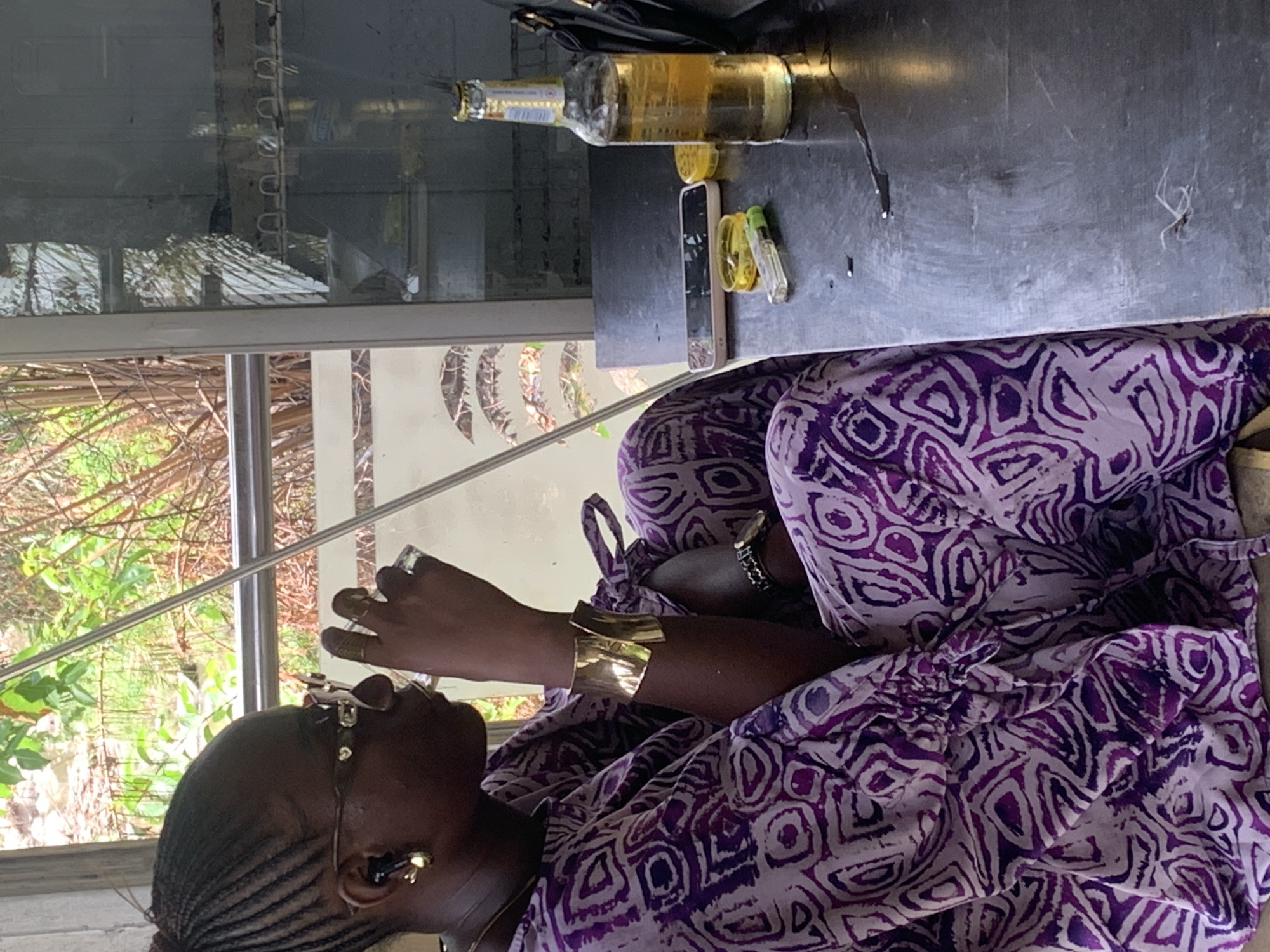 An African woman wearing glasses, earrings, and a large gold cuff bracelet, sits in what appears to be a bus or van. She wears a purple patterned dress. Outside the window is a view of green trees and foliage, suggesting a natural setting. On the table next to her is an open bottle of what appears to be beer, a smartphone, and some other small objects. The location is likely Adankolo, Nigeria.