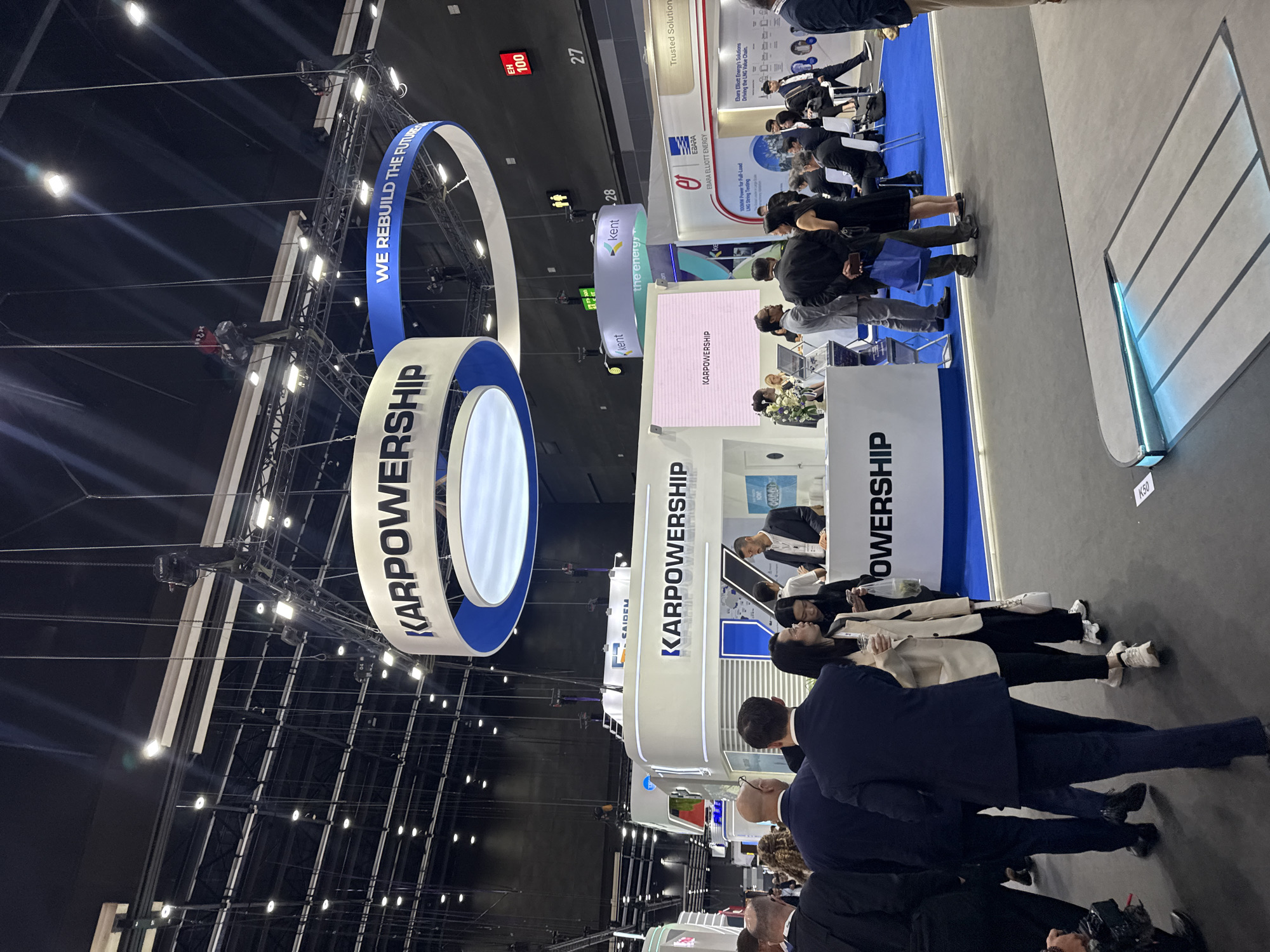 The image captures an indoor exhibition hall, likely at the IMPACT Exhibition and Convention Center in Bangkok, Thailand, given the scale and common exhibition types. A large, circular banner with "KARPOWERSHIP" prominently displayed hangs from the ceiling. Below this, a booth with the same branding is set up, featuring a white, modern design with blue accents. Several people are gathered around the booth, some appearing to be attendees and others possibly staff.
In the foreground, a group of people in business attire are walking. One individual in a dark suit is seen from behind, and a woman in a light-colored blazer and dark pants is walking beside him, holding a drink and a phone. The floor of the exhibition hall is a dark grey, with illuminated floor markings near the foreground.
In the background, other exhibition booths are visible, including one with the branding "Ebara Elliott Energy" and another featuring "Kent." Signs indicating booth numbers (27, 28) and directional information for restrooms are present on the ceiling infrastructure. The overall environment suggests a professional trade show or conference.