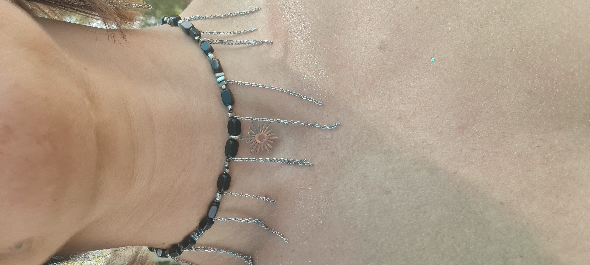 A close-up, angled shot shows a person's neck and upper chest. The person is wearing a necklace with dark stones and silver chains dangling from it. One of the chains has a sunburst charm. The skin on the neck and chest is light-toned and textured. A possible location of Lachish Regional Council, Israel cannot be determined.