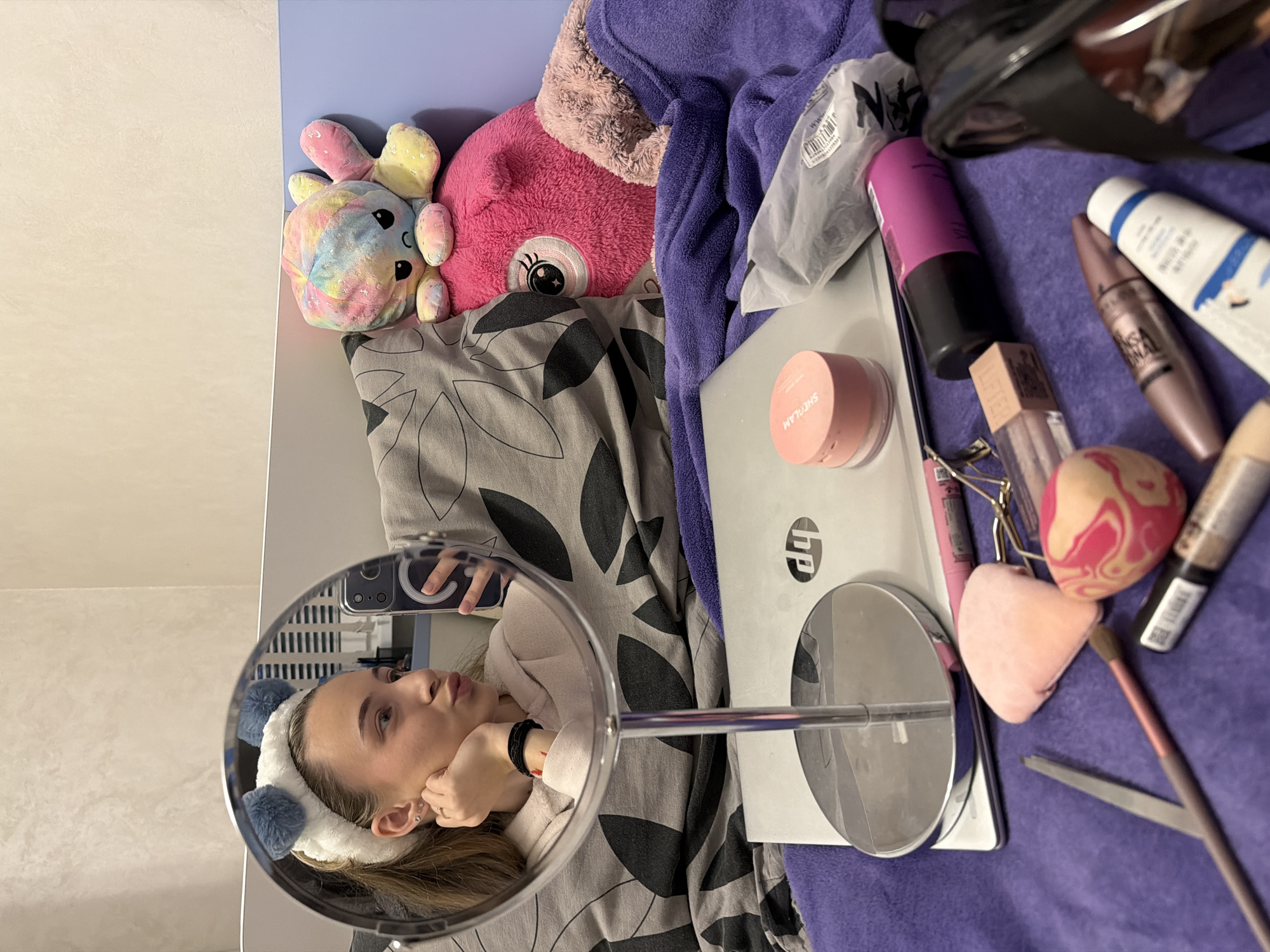The scene depicts an indoor environment, stated to be in Plovdiv, Bulgaria, specifically a bedroom or similar personal space. A young female is reflected in a round, silver-framed vanity mirror, positioned on a bed. She wears a white fluffy headband with two blue pom-poms, a light-colored hooded top, and holds a smartphone with a black magnetic ring holder in her left hand, which is also visible in the reflection. She is making a puckered or "fish-face" expression. Her right hand is near her jawline, displaying multiple ear piercings and a ring. A black elastic bracelet is visible on her left wrist.
The mirror's stand rests on a silver HP laptop, which is closed. The laptop and surrounding items are situated on a textured purple blanket or throw. To the right of the laptop, various cosmetic items are scattered: a pink Sheglam powder compact, several makeup brushes, pink and swirled pink/white makeup sponges, an eyelash curler, tweezers, mascara tubes (one identifiable as Maybelline), a pink cylindrical object, and other makeup containers. A white plastic bag containing an unknown item is also present.
Behind the laptop and cosmetics, a grey pillow with a repeating black leaf-like pattern is visible, alongside two plush toys. One is a reversible plush octopus, displaying its rainbow-colored "angry" side. Below it is a larger pink plush octopus with a single prominent eye. A light-colored wall, possibly beige or off-white, forms the background, with a segment of a light blue bed frame or headboard visible to the right.
