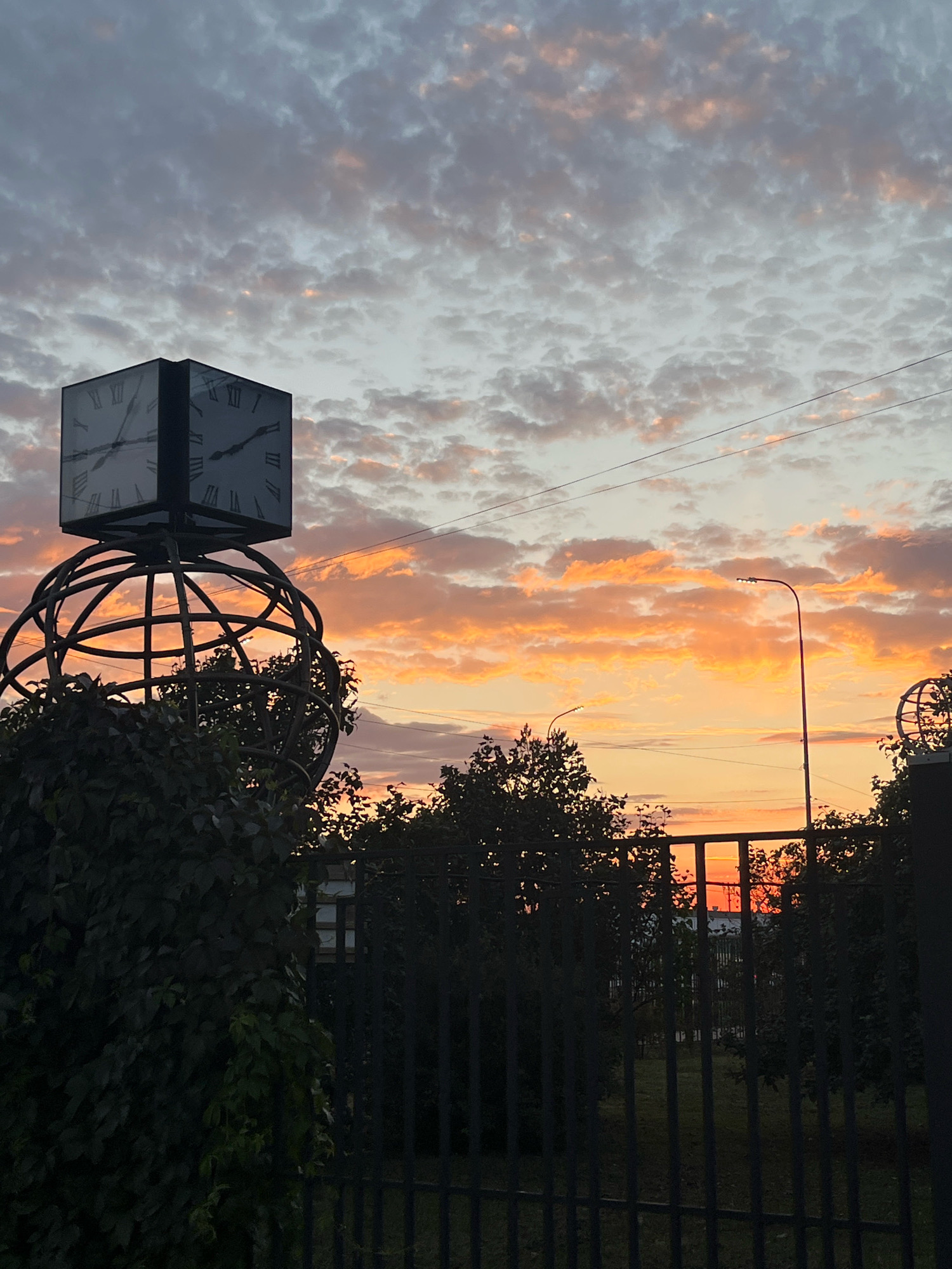 The image depicts an outdoor scene featuring a large, cubic clock with Roman numerals positioned atop an ornate, spherical metal structure. The clock is silhouetted against a sky filled with scattered clouds illuminated by the warm hues of a sunset, ranging from orange to pink. Green foliage obscures the lower part of the spherical structure. In the middle ground, a dark metal fence runs horizontally across the frame, with trees and a street lamp visible behind it. The background reveals more trees and buildings. A power line stretches across the upper part of the sky. The scene appears to be set in an urban environment, possibly Chaikovsky, Russia, given the context provided. There are no people or animals visible in the image, and no discernable actions are taking place. The overall impression is a quiet, late-day setting.