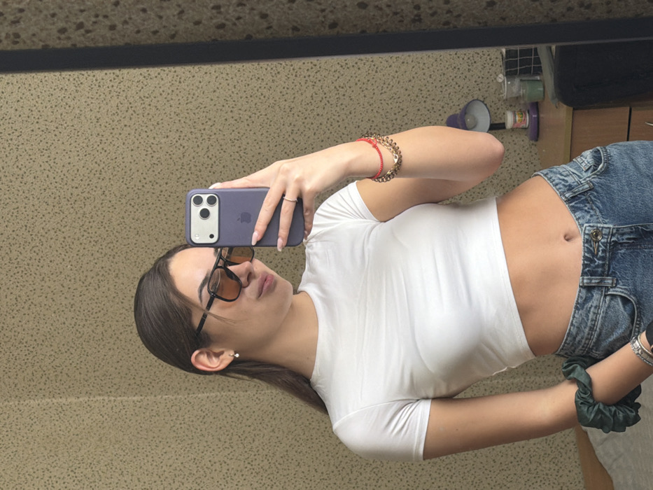 A young female subject is depicted taking a mirror selfie indoors. She has dark hair, wears dark-framed sunglasses with amber-tinted lenses, a white short-sleeved crop top, and light-wash denim jeans. A small drop earring is visible in her left ear. Her right wrist features multiple bracelets, including a red string and gold-colored chains, while a dark green scrunchie is on her left wrist. A ring is visible on her left ring finger. She holds a purple-cased smartphone, identified as an iPhone 14 Pro/Pro Max model, with her right hand, pointing it towards the mirror. The background, visible in the mirror's reflection, consists of a light brown/beige wall with a speckled texture. To the right of the subject, a purple desk lamp, various small containers, and a black grid organizer are present on a wooden desk. A portion of a bed or mattress with a light-colored sheet is reflected to the left. The specific location within Sofia, Bulgaria, cannot be determined from the visual evidence, as no identifiable external landmarks or unique interior features are present; the scene depicts a generic indoor room.