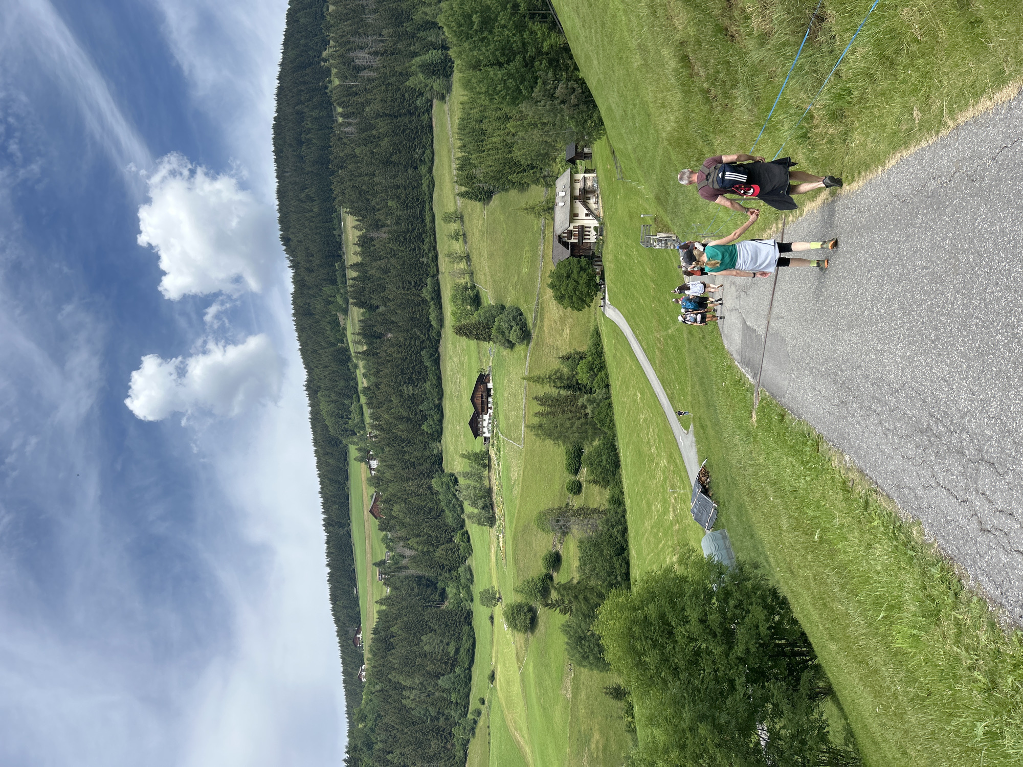 In an outdoor, rural landscape, a group of people walks along a paved path that winds uphill. The path is bordered by lush green grass and a forest. In the foreground, two individuals, a man and a woman, walk hand-in-hand, both wearing backpacks. The woman is in a teal tank top and grey skirt, and the man is in a red shirt and black jacket. Behind them, about seven other individuals walk together. Further up the path, scattered houses and more dense forest are visible on rolling green hills. The sky above is partly cloudy with scattered white clouds against a blue sky. The scene appears to be a scenic hiking trail.