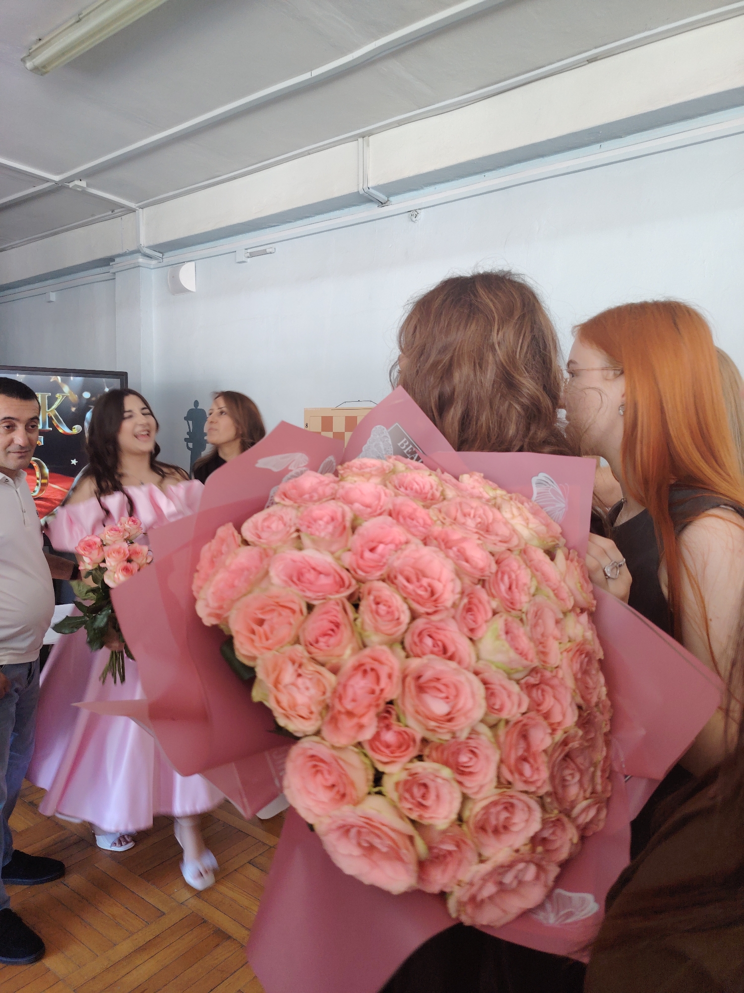 This image captures a celebratory moment, likely a graduation or special occasion, taking place indoors in Rostov-on-Don, Russia. The central focus is a young woman in a light pink, off-the-shoulder dress, beaming with joy and laughter. She is holding a large bouquet of pale pink roses, with another bouquet visible in the foreground.
To her left stands a man in a white polo shirt and blue jeans, looking towards the woman with a subtle smile. Behind the young woman, a woman with long brown hair is partially obscured, and to the right, another woman with long, reddish-orange hair and glasses is leaning in, seemingly whispering or sharing a moment with the woman holding the bouquet. The interaction between the people suggests a warm and affectionate atmosphere.
The setting appears to be a room with light-colored walls and a white ceiling with fluorescent lighting. A wooden floor with a parquet pattern is visible. In the background, a large screen displays some text that includes the letter "K" in a decorative font. The overall lighting suggests it is daytime, though the specific weather conditions are not discernible. The presence of bouquets of roses and the celebratory mood of the people point towards a happy event.