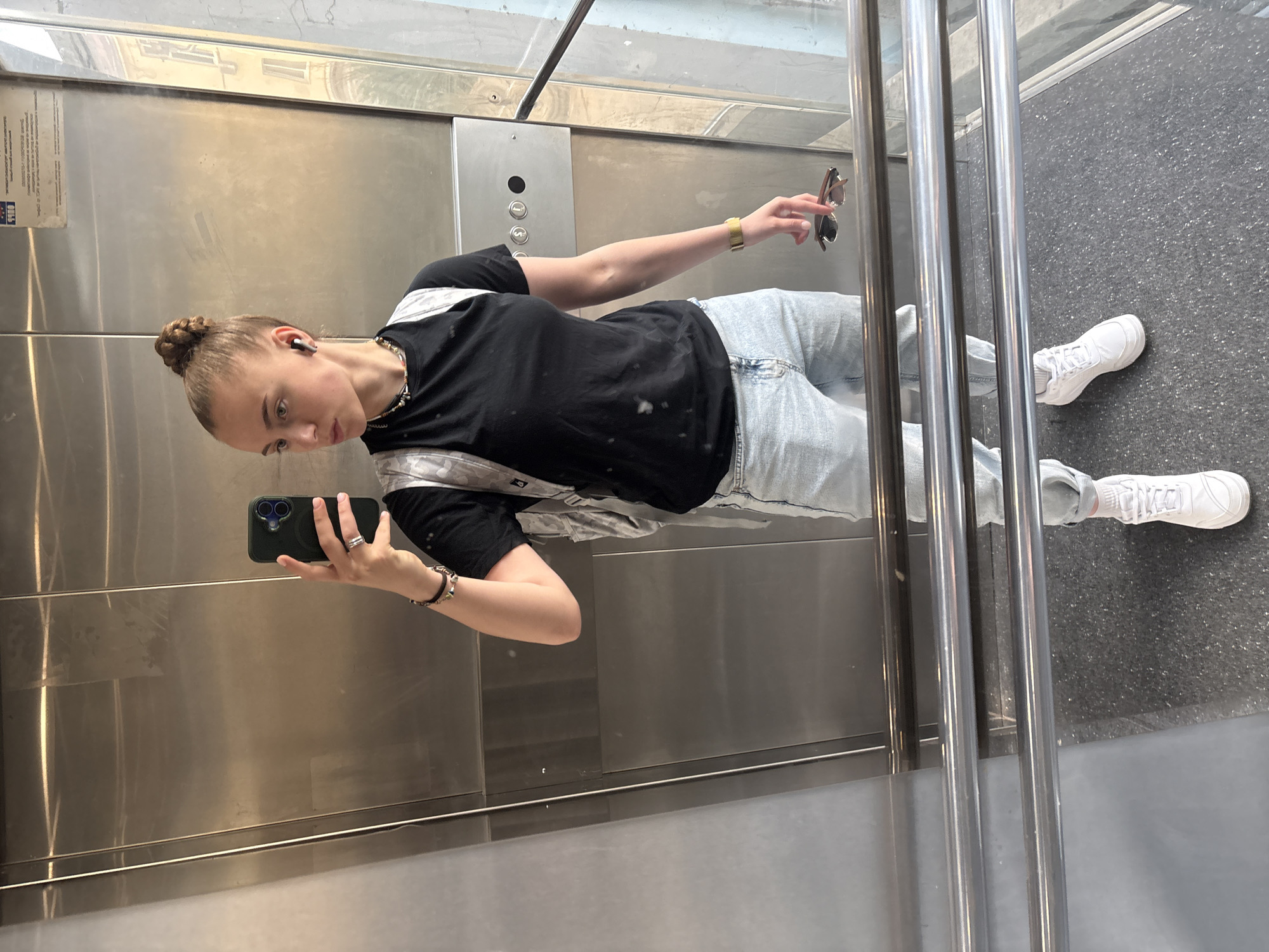 A young woman is taking a selfie in an elevator. She has her hair in a bun and is wearing a black T-shirt, light wash jeans, and white sneakers. She is holding a dark-colored smartphone in her left hand and sunglasses in her right. The elevator has stainless steel walls and a control panel with buttons. The floor of the elevator is dark gray with a speckled pattern. The location is likely Sofia, Bulgaria, due to details visible in the reflections of the metal, the style of elevators, and the type of buildings near the elevator.