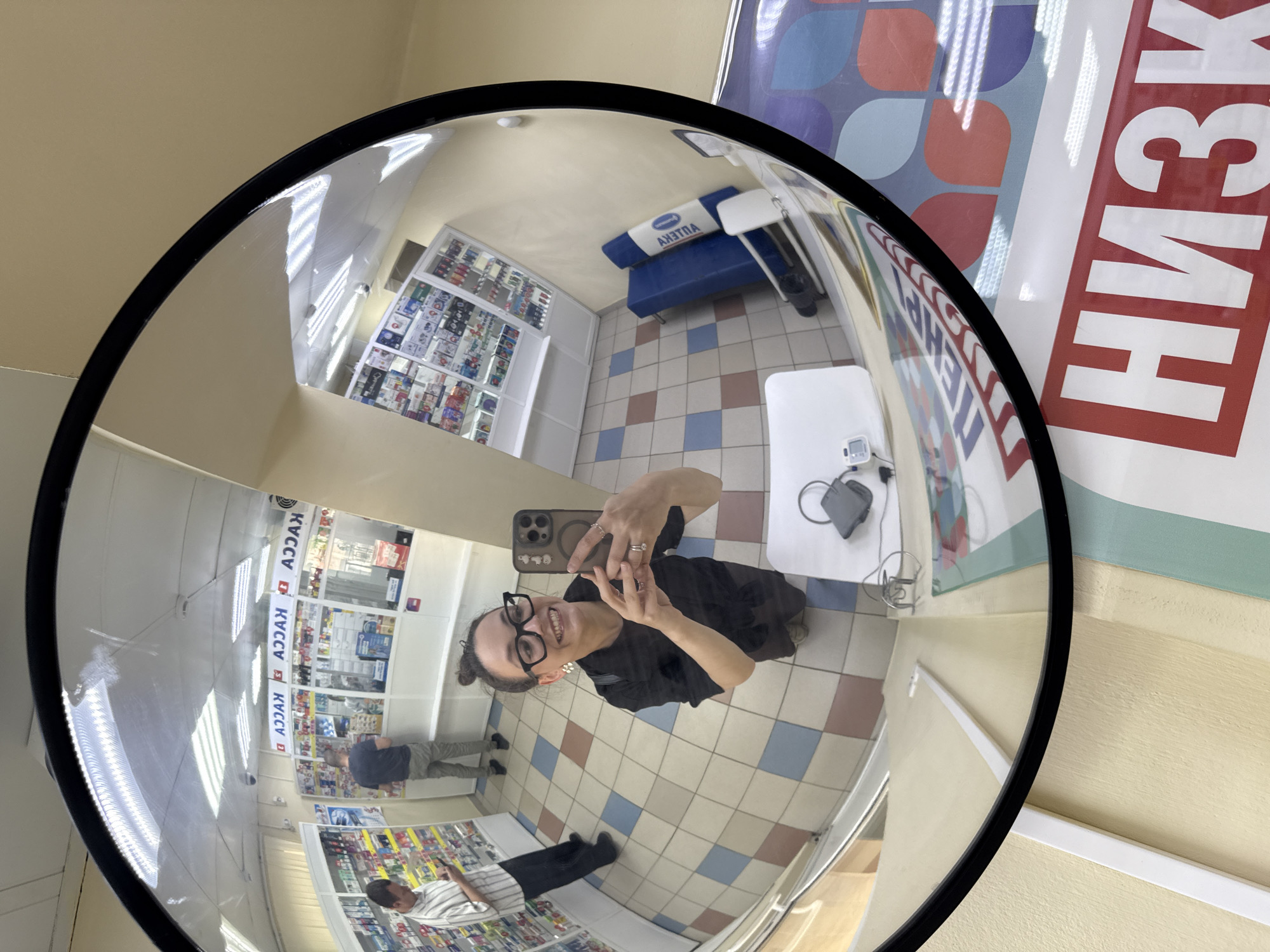 The image is a fisheye mirror selfie taken inside a pharmacy in Chaikovsky, Russia. A woman with dark hair pulled up in a bun, wearing black clothing and glasses, is smiling and holding a smartphone, taking the picture. She is wearing rings on her fingers and white earrings. In the background, there are shelves stocked with various products, and a man in a striped shirt and dark pants is looking at his phone. Another man in a dark t-shirt and grey pants is standing near a counter. The pharmacy has a tiled floor with a pattern of beige, red, and blue squares. A blue bench is visible in the corner of the store. Above the counter, a sign with Cyrillic text, "КАССА" (KASSA - meaning "cashier" or "checkout"), is reflected in the mirror. A large banner with the Cyrillic word "НИЗК" (NIZK - likely part of "НИЗКИЕ ЦЕНЫ" meaning "low prices") is visible on the right side of the image. The overall environment appears to be a retail space.