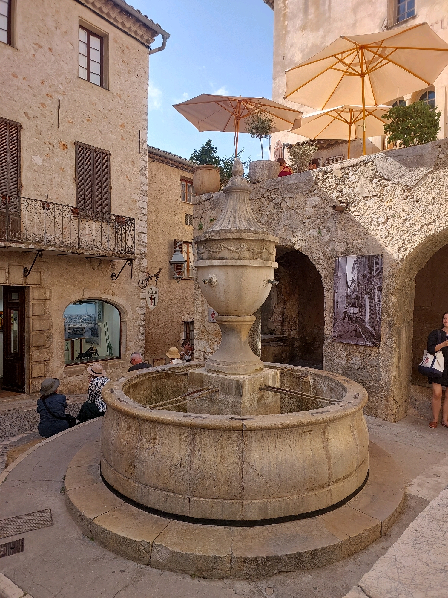 The image depicts a sunlit, ancient stone fountain in the center of a narrow, cobbled street in the medieval village of Saint-Paul-de-Vence, France. The fountain is substantial, with a large, tiered basin and an ornate central structure topped with a decorative finial. Water spouts are visible on the fountain.
To the left of the fountain, a two-story building with a rustic, textured stone facade features wooden shutters, a wrought-iron balcony, and a shop entrance. The window of the shop displays a miniature diorama.
To the right and behind the fountain, an arched passageway leads into a shaded area. A large black and white photograph of a narrow street scene is mounted on the stone wall within this archway. A woman in a dark dress and holding a white tote bag stands to the right of the archway.
Several people are present in the scene:
* Two individuals, viewed from the back, are seated on the edge of the fountain in the lower-left quadrant of the image, wearing hats.
* A group of people are seated at a table under umbrellas on a raised stone terrace in the upper-right background.
* A person is partially visible sitting on the terrace, wearing a red top.
Overhead, large cream-colored umbrellas provide shade for an outdoor seating area on the elevated terrace. The architecture is consistent with the historic character of Saint-Paul-de-Vence, characterized by its stone buildings, narrow streets, and open courtyards. The overall atmosphere is that of a charming, well-preserved European village on a clear day.