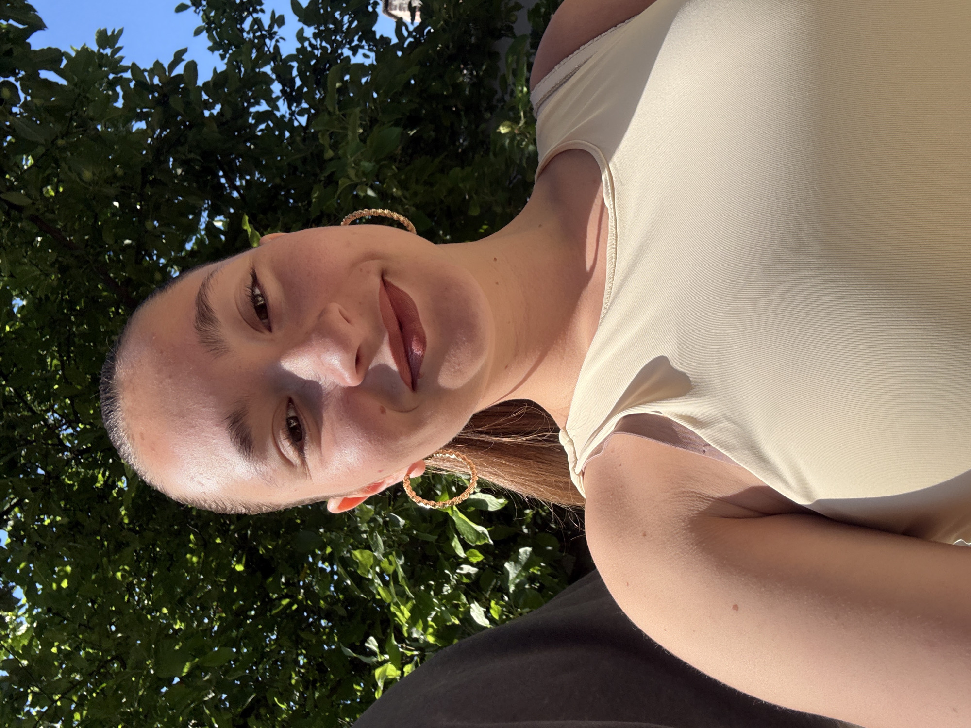 A young woman with light brown hair pulled back into a ponytail and wearing large hoop earrings is captured in a close-up selfie. She is smiling, with her lips painted in a dark berry shade. The woman is wearing a sleeveless, light beige ribbed top. She is outdoors, with a backdrop of lush green foliage from a tree and a clear blue sky. The sunlight creates a warm glow on her skin. The image was taken at Vitosha Mountain, a popular recreational area on the outskirts of Sofia, Bulgaria.