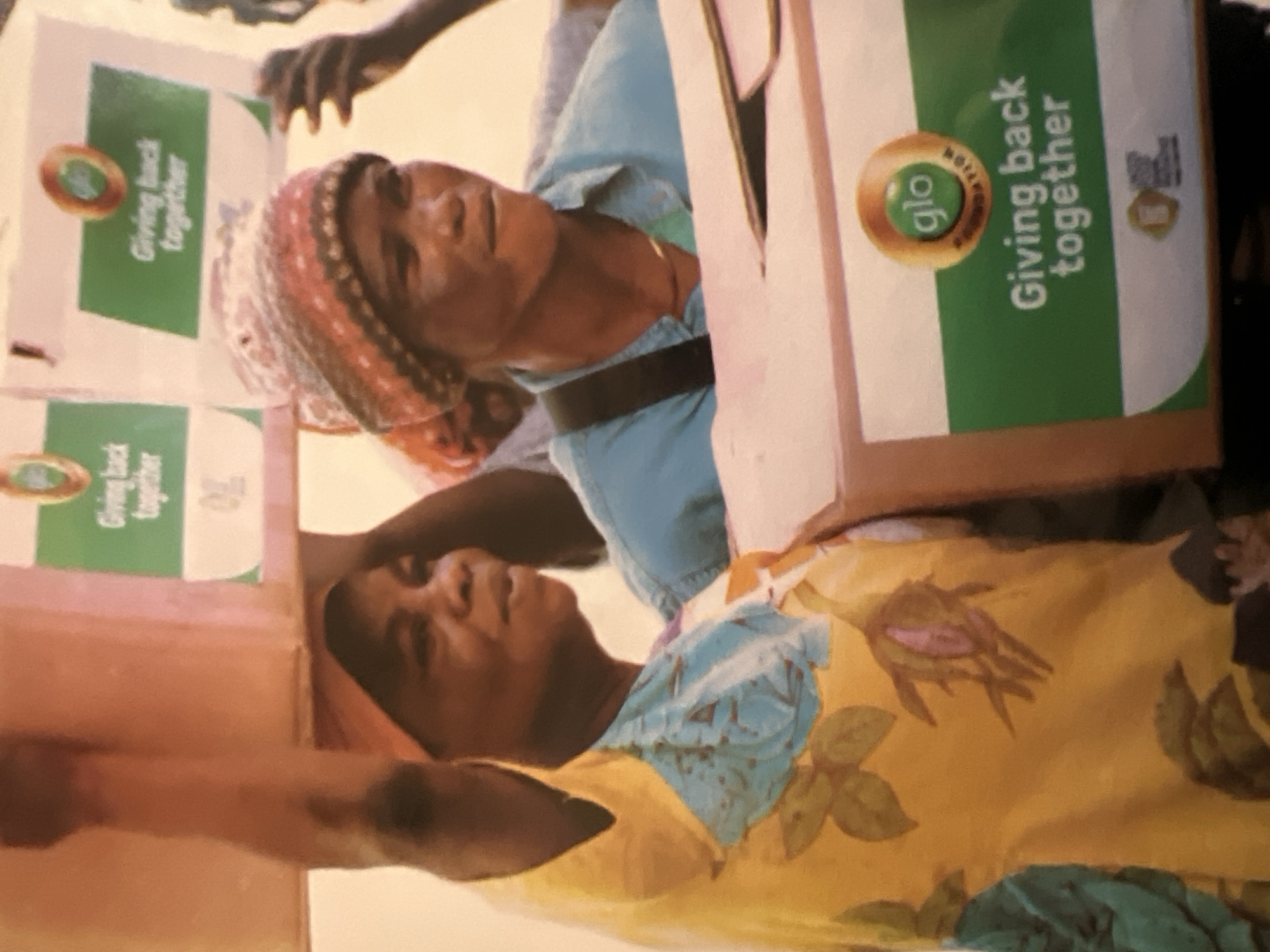 In Jalingo, Nigeria, two women are seen carrying large cardboard boxes. The woman on the left, wearing a yellow and blue floral top, balances a pink box on her head. The woman on the right, with a knitted hat, holds a box with green and white branding that reads "Glo Foundation: Giving back together." The background is indistinct, suggesting an outdoor or open indoor setting.