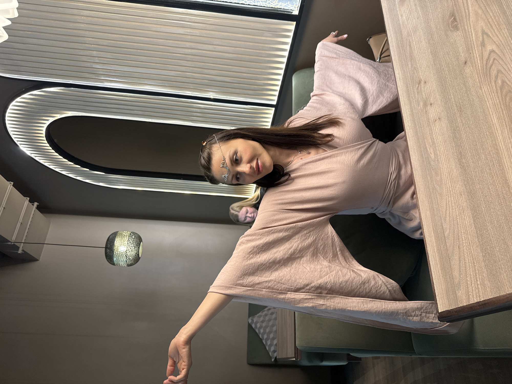 A young woman with long brown hair is sitting at a table in what appears to be a cafe or restaurant. She is wearing a light pink robe-like garment with wide sleeves and a decorative headpiece. The room is modern, with a curved, lit ceiling feature, a spherical light fixture, and a wooden table. Another person with blonde hair is partially visible in the background. The location is Unknown, Russia.