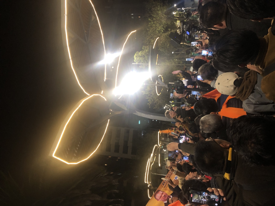 A crowd gathers at night under a large, brightly lit structure resembling stylized leaves. Many people are recording the event with their phones. The light from the structure illuminates the dark sky. A sign with a man's portrait is visible among the crowd. Trees and buildings are visible in the background, with street lights further adding to the overall illumination of the scene.