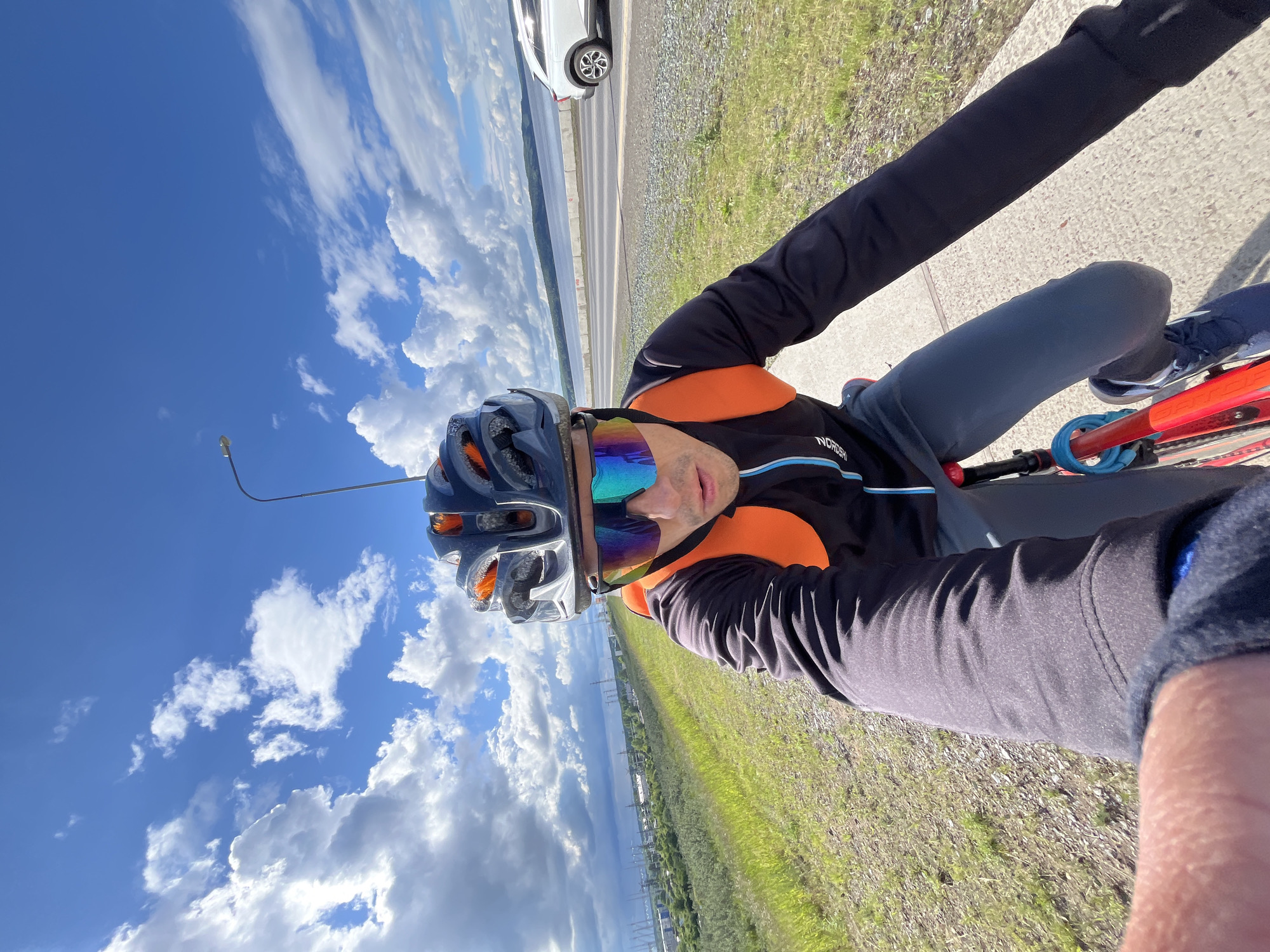 The image shows a first-person selfie perspective of a person on a bicycle, taken in Chaikovsky, Russia. The subject, an adult male, is wearing a dark grey cycling helmet, vibrant iridescent reflective cycling glasses, a black long-sleeved cycling jersey with orange accents, and grey cycling pants. Part of a red bicycle frame with a blue U-lock is visible. The jersey has "NORDSHI" printed on it.
The scene is outdoors on a bright, sunny day with a clear blue sky dotted with scattered white, fluffy clouds. The person is positioned on a paved road or path that runs alongside a wide body of water, likely a river or reservoir. A grassy shoulder is adjacent to the road. In the background, a white car is visible on the road, and across the water, distant landmasses and structures, possibly including power lines, can be seen. A street light pole extends into the sky on the left.
The activity depicted is cycling or a pause during a bike ride. The bright daylight suggests it is midday or early afternoon, with excellent weather conditions for outdoor exercise. The overall impression is of an active individual enjoying a scenic ride.