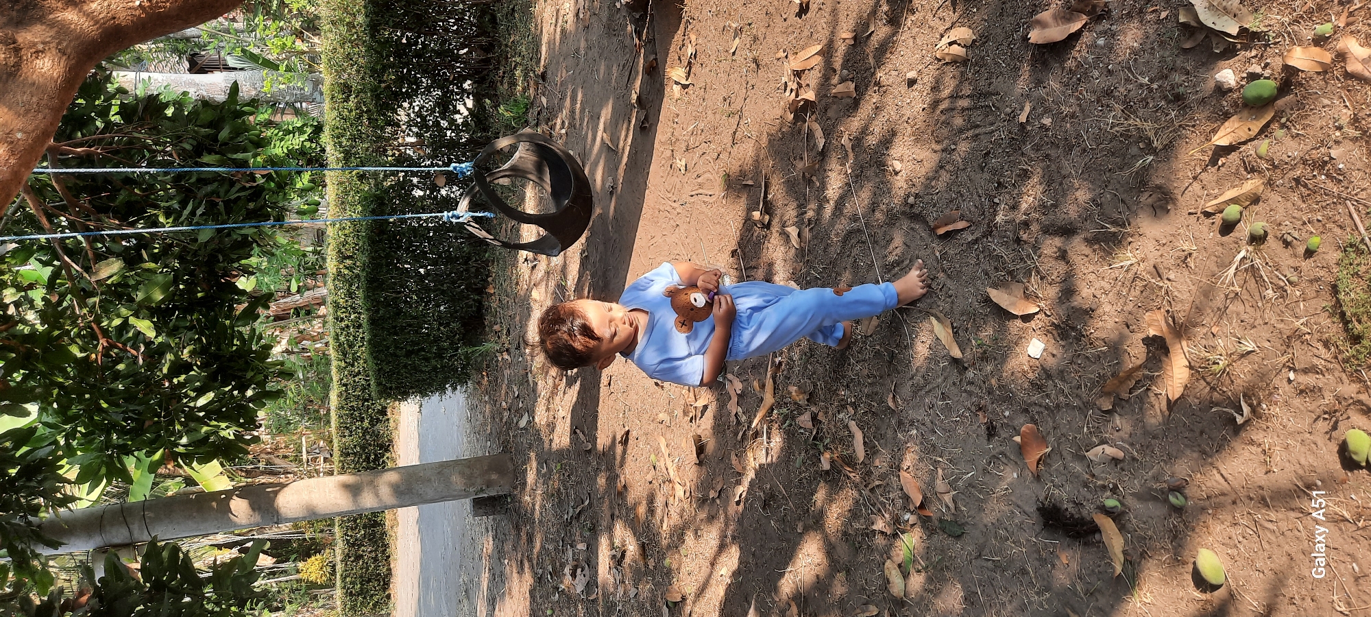 A young child, wearing light blue pajamas with a teddy bear applique, stands barefoot on a dirt ground covered with fallen leaves and scattered green mangoes. The child is holding a small brown teddy bear and looking down. Behind the child, a black tire swing hangs from a tree. In the background, a neatly trimmed green hedge runs along the edge of a paved area. The scene is lit by sunlight, casting dappled shadows from the surrounding trees. The location is Ploso, Indonesia.