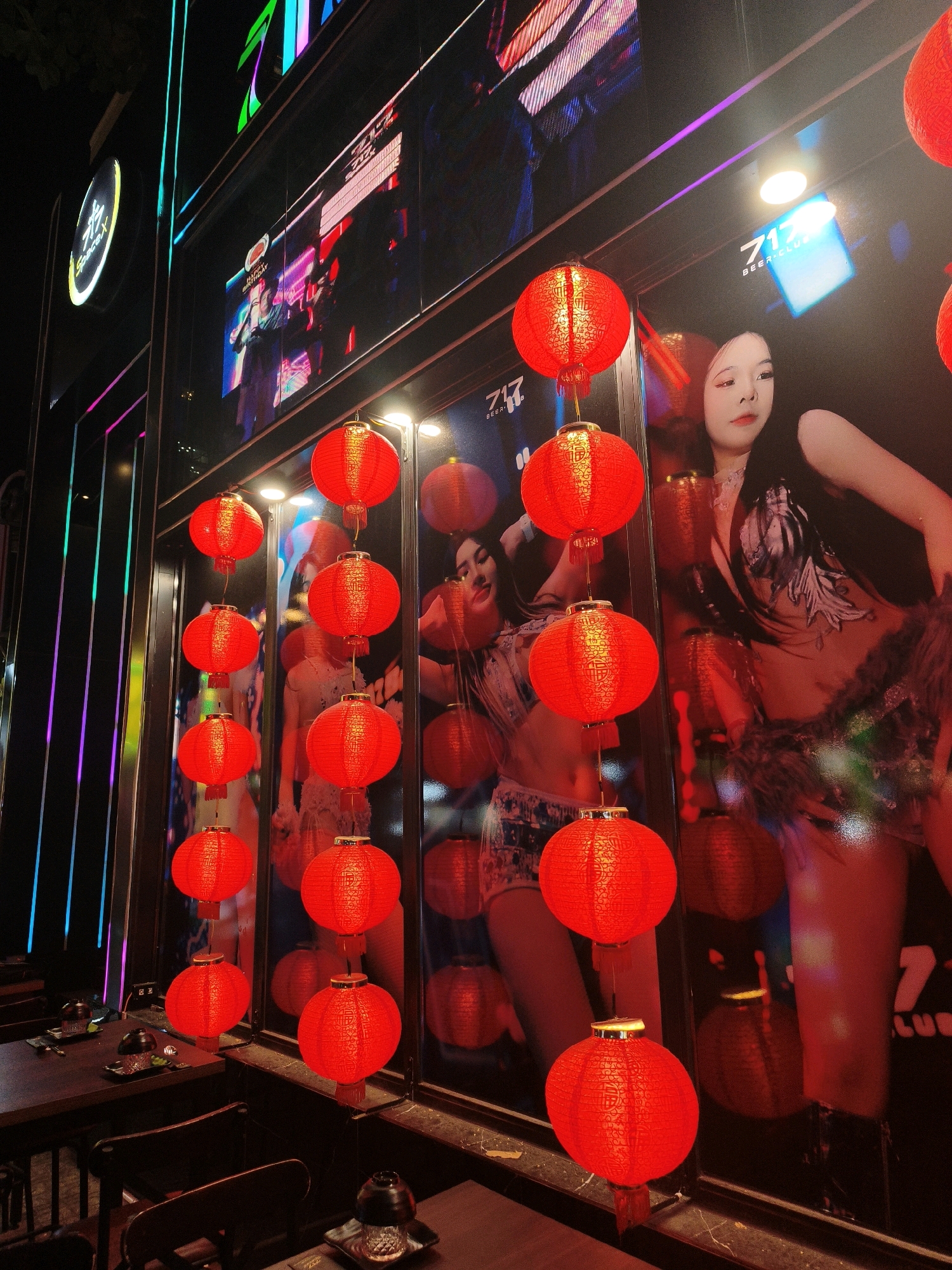 The image captures a nighttime scene in Nha Trang, Vietnam, featuring the exterior of a brightly lit establishment called "717 BEER CLUB." The facade is adorned with large, illuminated digital screens displaying vibrant graphics, including advertisements and images of women. Hung in front of the screens are numerous traditional red lanterns, some with intricate patterns and tassels, casting a warm glow. The foreground shows a section of outdoor seating with dark wooden tables and chairs, set for patrons. The overall atmosphere is lively and commercial, typical of entertainment venues in urban areas.