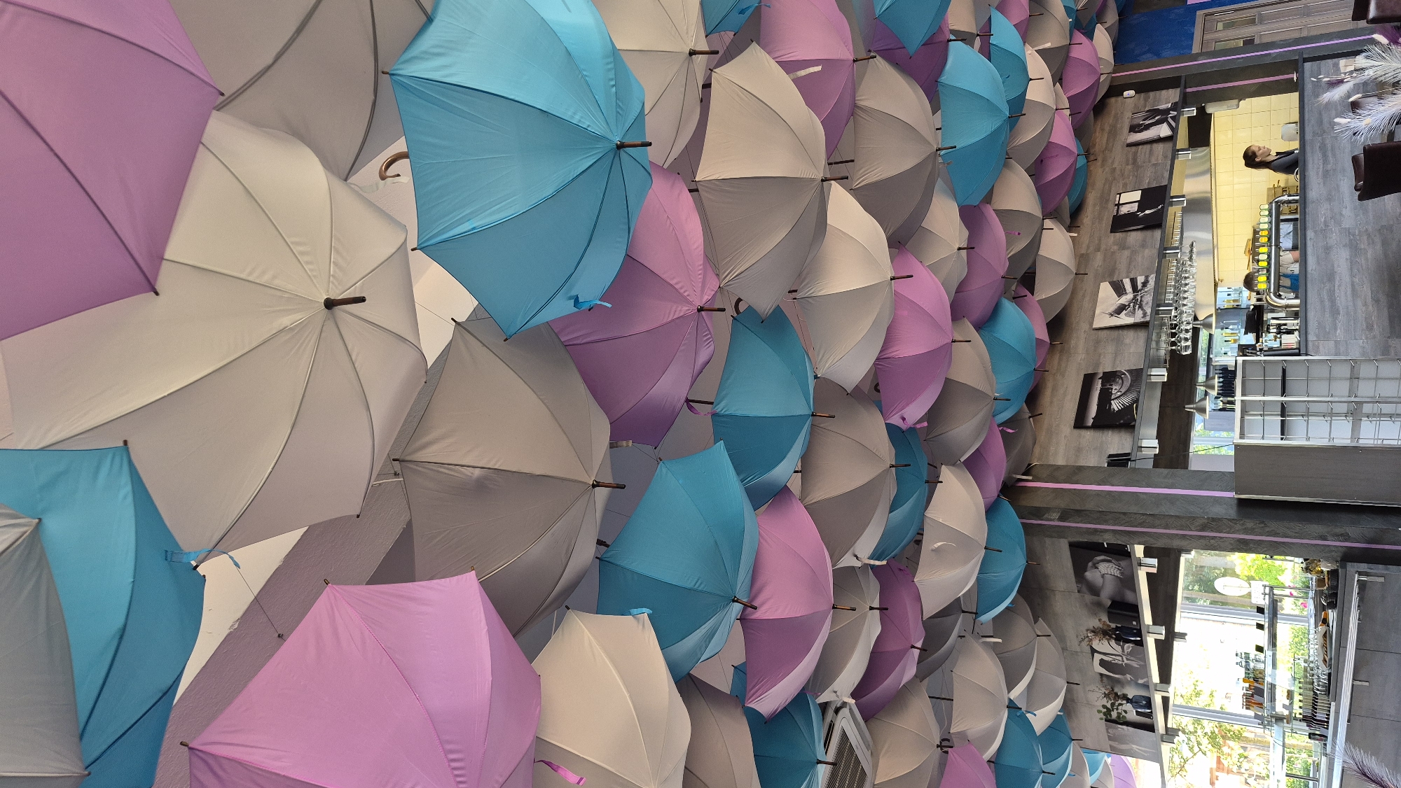 The image depicts the interior of a cafe or restaurant, possibly located in Zarasai, Lithuania, as suggested by the prompt. The ceiling is adorned with a dense arrangement of upside-down open umbrellas in various shades of pink, light blue, and gray. Below the umbrellas, the environment appears to be a functional dining or service area.
The upper wall features a shelf displaying black and white framed photographs, with some floral arrangements. The main area below includes a counter with a visible coffee machine and bar equipment, where two individuals, likely staff members, are present. One individual, a woman, is behind the counter, interacting with equipment. Another person, partially visible, is also behind the counter.
In the background, a window offers a glimpse of outdoor greenery, suggesting a connection to the outside. The foreground and midground show more of the cafe's interior, with tables and chairs, and shelving units. The overall impression is of a decorative and perhaps whimsical interior design, juxtaposed with the practicalities of a food and beverage establishment.