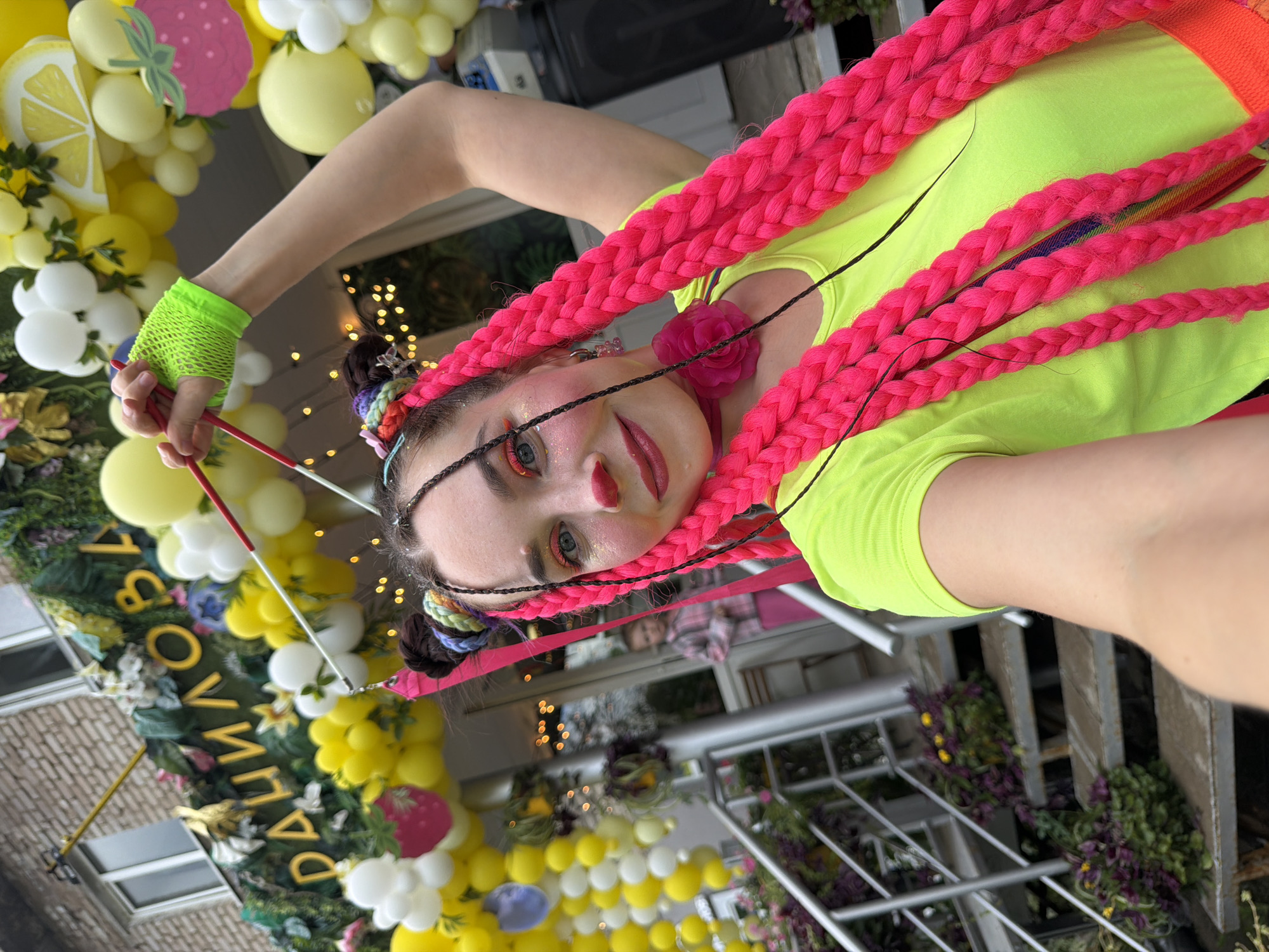 A young woman with brightly colored braids and clown makeup is taking a selfie in front of a building decorated with balloons, leaves, and large yellow letters spelling "ЧАЙКОВСКОЙ" (Chaikovskoy), a clear reference to the city of Chaikovsky, Russia. She is wearing a neon green t-shirt and holding a red stick. The background shows a building entrance with stairs and greenery, and a child in a pink shirt is visible in the doorway. The overall scene suggests a festive or celebratory event.
