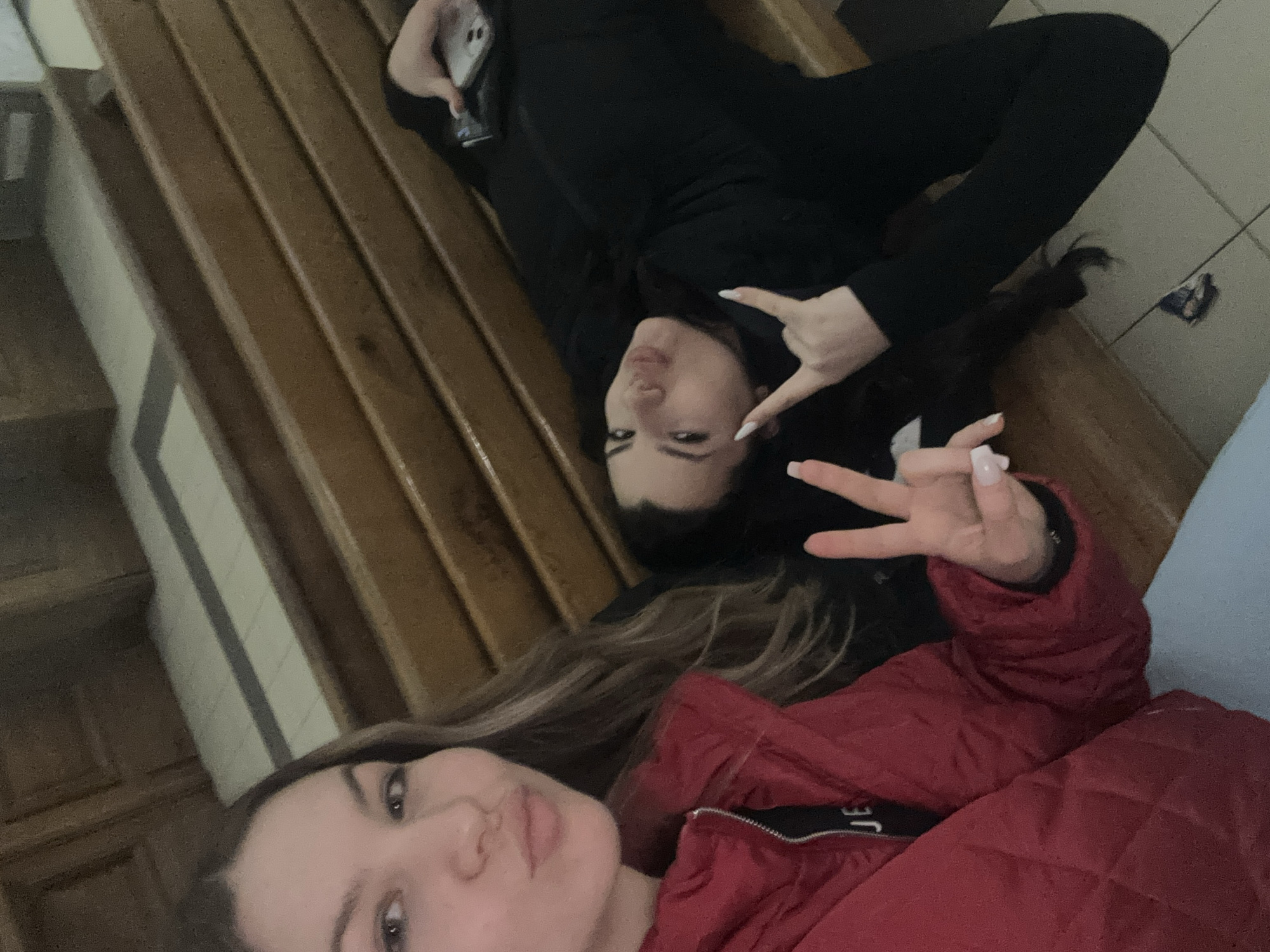 Two young women are lying down on a wooden bench. One woman is wearing black clothing, and she's holding a mobile phone. The other woman is wearing a red jacket. Both women are making a V-sign with their fingers. The bench appears to be located indoors, possibly in a waiting area or lobby. The background suggests a public space with tile flooring. It's not possible to verify a location in Burgas, Bulgaria, from the image provided.