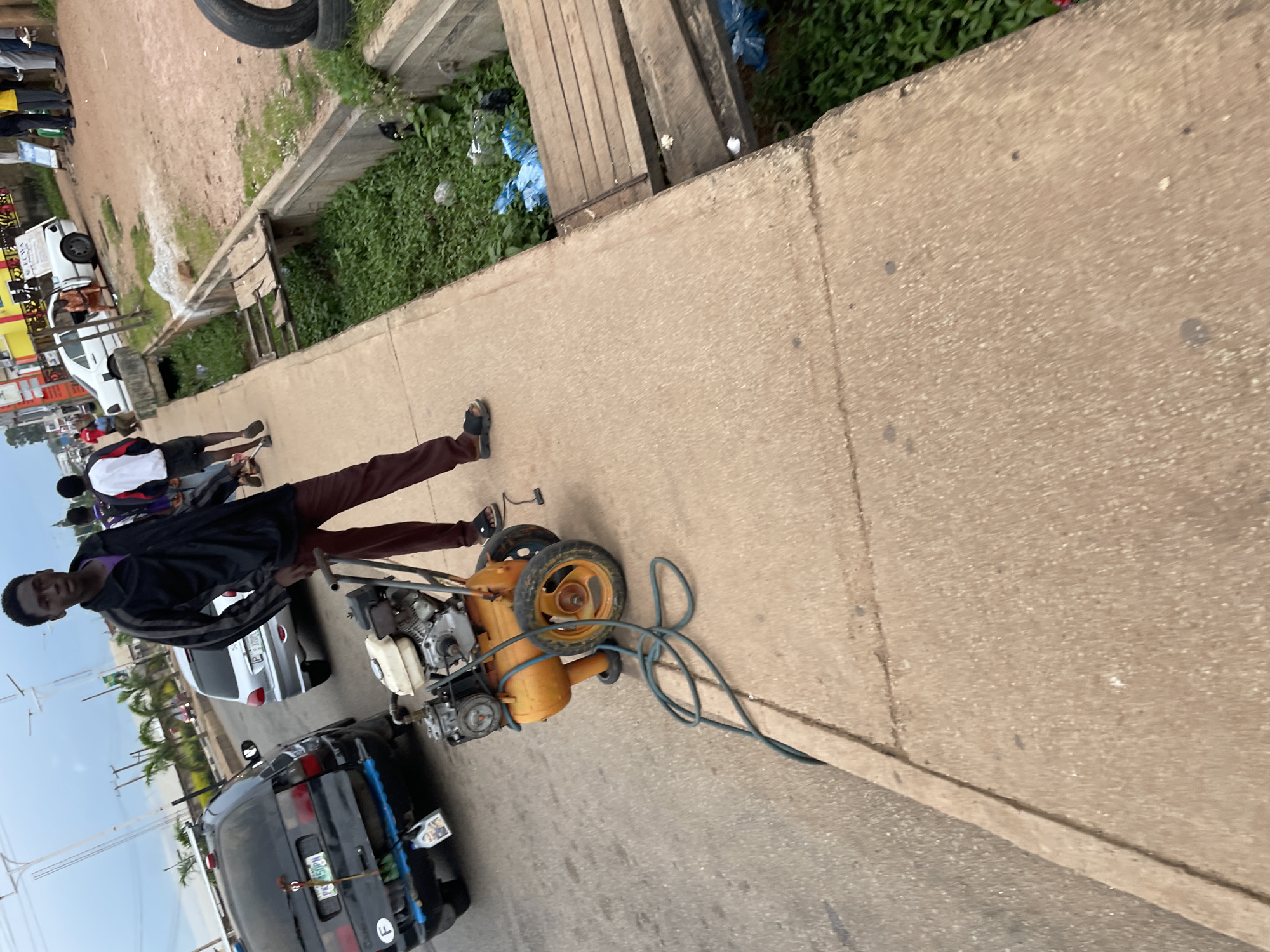 A young Black male, wearing a black jacket over a purple shirt and maroon pants, stands on a concrete sidewalk next to a small, yellow air compressor with two wheels. The air compressor has a gas engine and a coiled green hose. In the background, traffic moves along a paved road. A black SUV is parked on the left, and a silver hatchback is driving away. Further down the road, a white car is parked and several people stand around it. A drainage ditch with green foliage and blue plastic bags runs alongside the sidewalk. The scene appears to be in Jos, Nigeria.