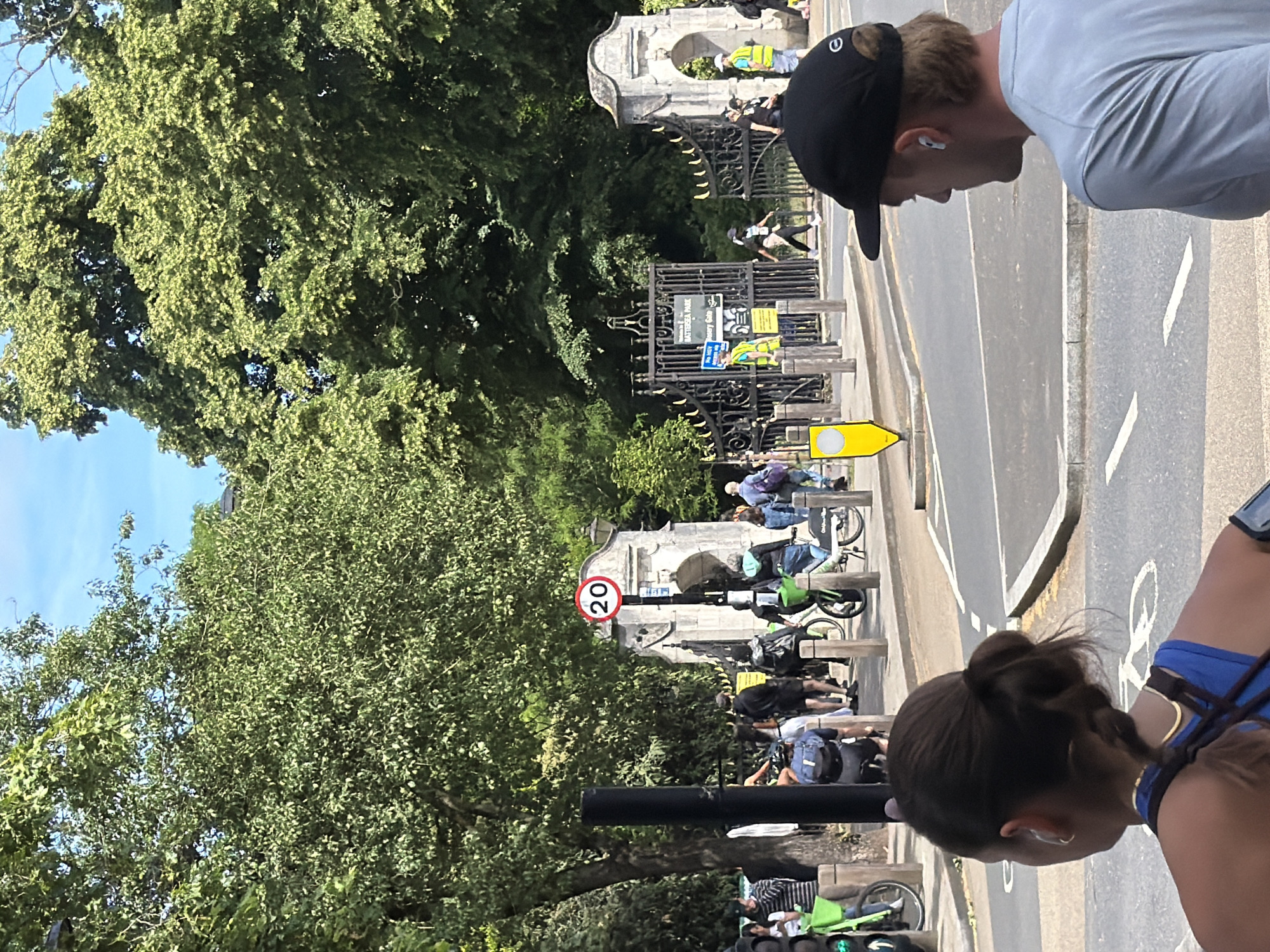 The image shows an outdoor scene with people and trees in the background. In the foreground, a woman with her hair in a bun is looking to the left, wearing a blue top and a gold necklace. To her right, a man wearing a black cap and a grey t-shirt with wireless earbuds in his ears is looking to the right. In the middle ground, there is a park entrance with a black metal gate and a stone archway. Several people are seen walking and cycling through the gate. One person is wearing a high-visibility vest and holding an umbrella. A speed limit sign showing "20" is visible on a pole near the entrance. The environment appears to be a park in London, United Kingdom, possibly Battersea Park, given the sign on the gate. The weather is sunny with a blue sky and green trees.