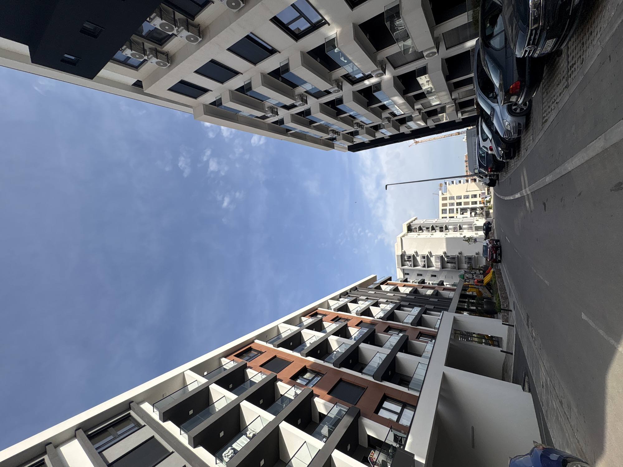 The image depicts a street view in Bar, Montenegro, taken from a low angle looking upwards. Two modern apartment buildings flank the street, creating a narrow corridor of sky. The building on the left has a predominantly white facade with alternating sections of brown panels and dark-framed windows and balconies. The building on the right features a white facade with dark-framed windows and balconies, with numerous air conditioning units. Parked cars line the right side of the street, partially obstructing the view of the building facades further down. In the distance, other buildings are visible under a blue, slightly cloudy sky. A lamppost stands tall near the center of the image, in line with the street. A glimpse of a playground can be seen between the buildings on the left side of the street.