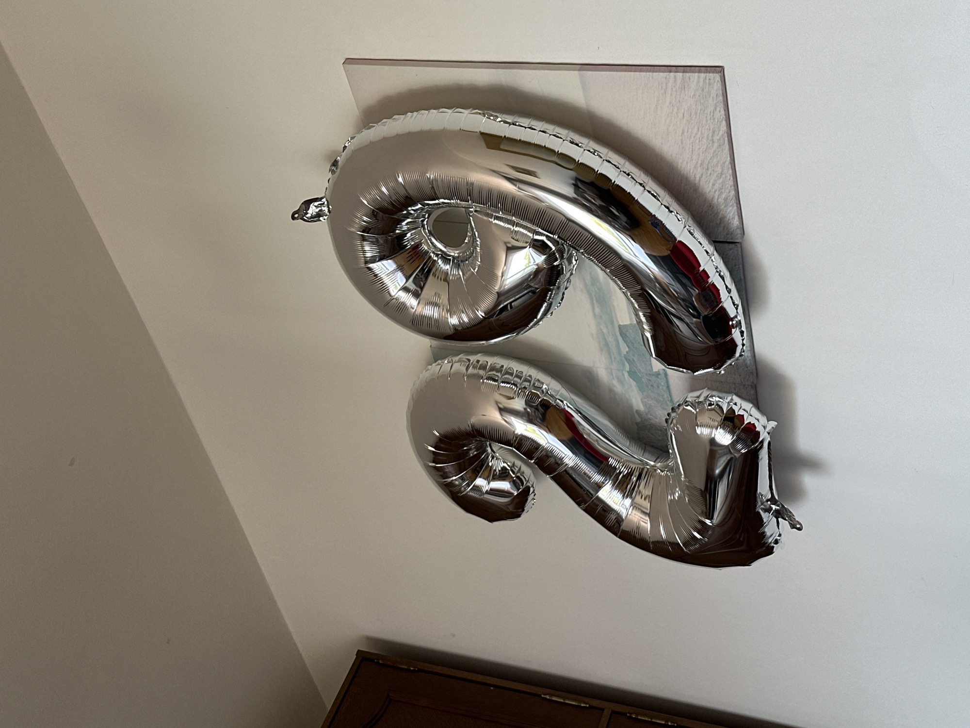 The image depicts two large, silver foil balloons, shaped as the numbers '2' and '9', attached to a white wall. The balloons are reflective, showing distortions of the surrounding environment. Behind the balloons, a framed artwork with a landscape scene of water and distant land is partially visible. To the left, a dark wooden cabinet or door is present. The overall setting appears to be an indoor residential space. There is no indication of the location being in the city of Grad Rijeka, Croatia.