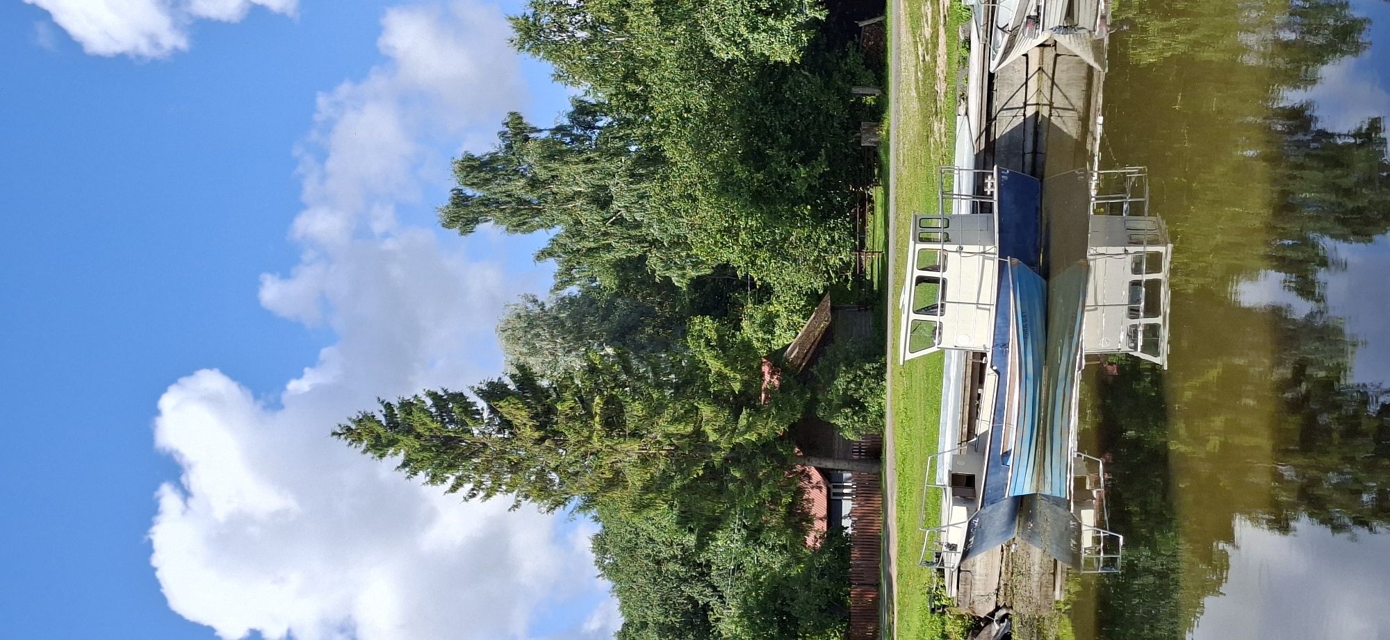 This is a vertical, eye-level, outdoor photograph of a waterfront scene in Valma, Estonia. The foreground features the tranquil, reflective surface of a body of water, likely a lake or river, with the mirrored image of the sky and surrounding trees. Moored to a concrete dock in the middle ground are two boats. The larger boat, to the right, is white with a blue hull and features a cabin with windows. Beside it, to the left, is a smaller, bright blue boat with a white interior and a railing. The background is dominated by lush, green trees and foliage, with a few wooden structures, possibly cabins or sheds, partially visible amongst the trees. A wooden fence runs along the edge of the property. In the distance, a tall evergreen tree stands out against a bright blue sky dotted with white, fluffy clouds. The overall impression is one of peaceful rural life by the water.