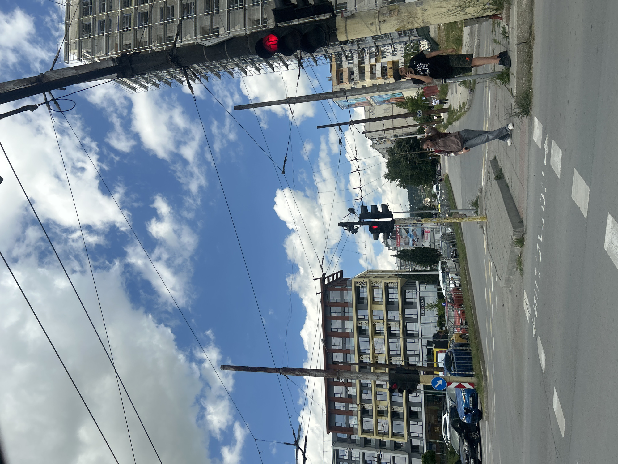 The image depicts a street scene in Varna, Bulgaria, likely at an intersection. The sky is blue with scattered white clouds. Overhead, a network of power lines and trolley wires crisscrosses the frame, supported by utility poles.
In the foreground, a street with lane markings and a pedestrian crossing is visible. Several cars are parked along the side of the road, and one blue car is in the lane. A traffic light showing red is prominently positioned to the right.
Two young people are standing on the sidewalk near the traffic light. One person, appearing to be a female, is wearing a light brown hooded sweatshirt and jeans and is looking towards the left. The other person, appearing to be a male, is wearing a black t-shirt and camouflage shorts and is holding a phone to his ear, looking towards the right.
Behind the people and the traffic light, several buildings are visible. To the left, a modern apartment building with balconies and large windows stands out. To the right, a taller building is under construction, covered in scaffolding. Other buildings, some appearing older and in various states of repair, line the street. Trees and vegetation are present on the sides of the road. Advertisements and signage are visible on some of the buildings and utility poles. The overall impression is a busy urban environment with ongoing development.