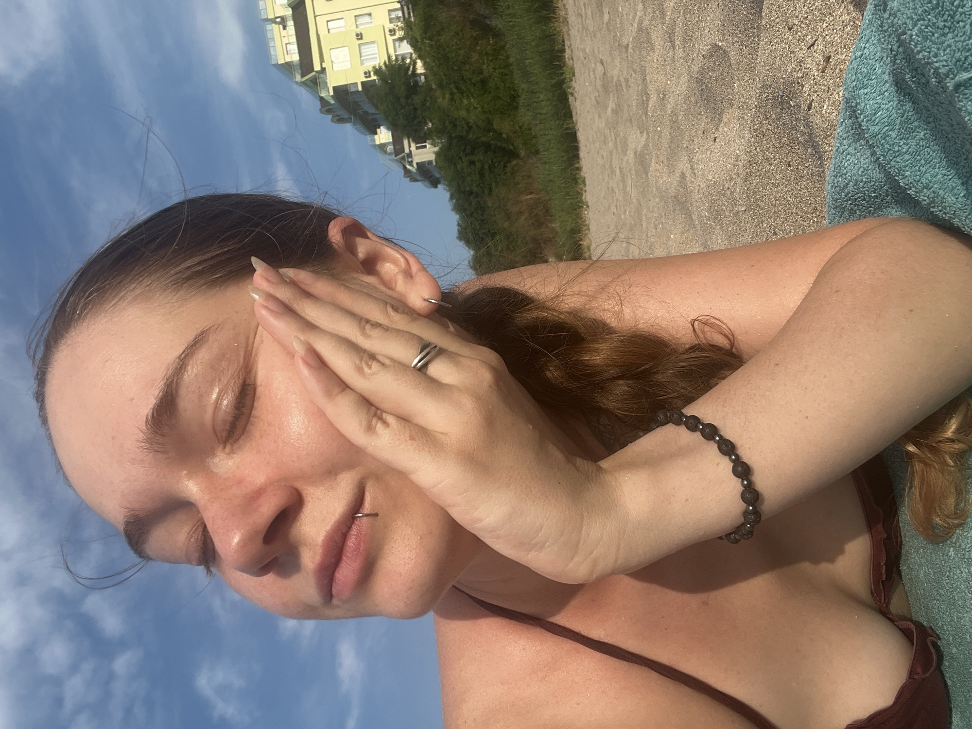 The image features a close-up of a young woman lying on a beach. She has her eyes closed and her hand resting on her cheek, with her fingers extended upwards. She is wearing a dark-colored bikini and a beaded bracelet on her wrist. Her hair, which appears to be reddish-brown, is partially visible and styled in a braid. A silver ring is on her ring finger, and she has a lip ring.
The setting is a beach, indicated by the sand beneath her and a teal towel to her right. In the background, there is a body of water that appears to be the sea, and behind that, a lush green hillside with trees. A modern yellow building with several windows is visible on the hillside, suggesting an urban or resort area nearby. The sky is blue with scattered white clouds, and the lighting suggests it is daytime, likely late afternoon or early evening due to the warm glow on her skin. The overall mood is relaxed and serene, with the woman seemingly enjoying the sun and the beach environment. There is no visible text in the image.