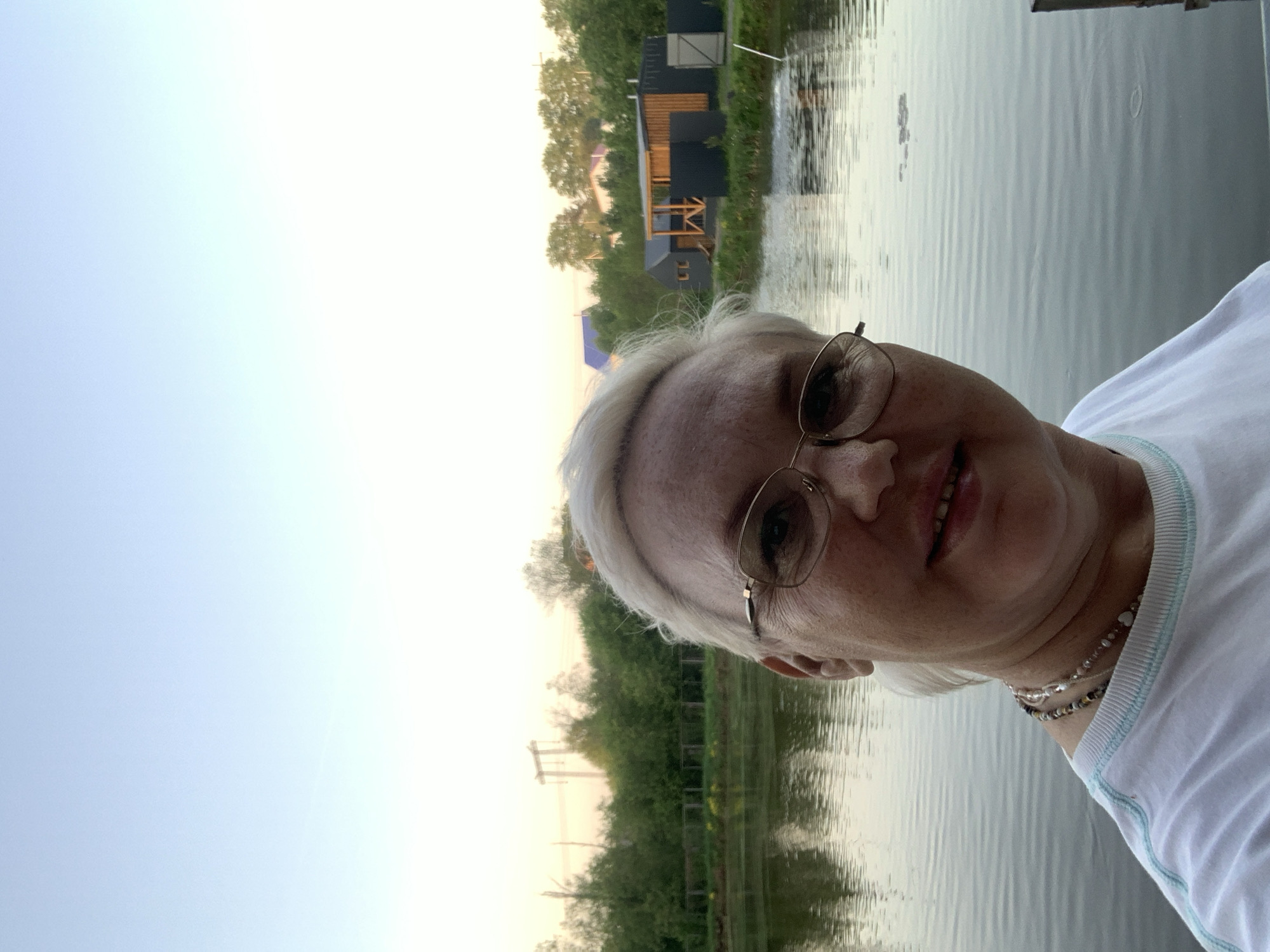 A selfie taken outdoors at twilight or dawn shows a blonde woman wearing glasses and a white t-shirt with a light blue trim, a beaded necklace. She is positioned with a body of water behind her, reflecting the sky. Beyond the water, there's a line of trees. To the left, a building. The location is most likely Malyi Bukor, Russia.